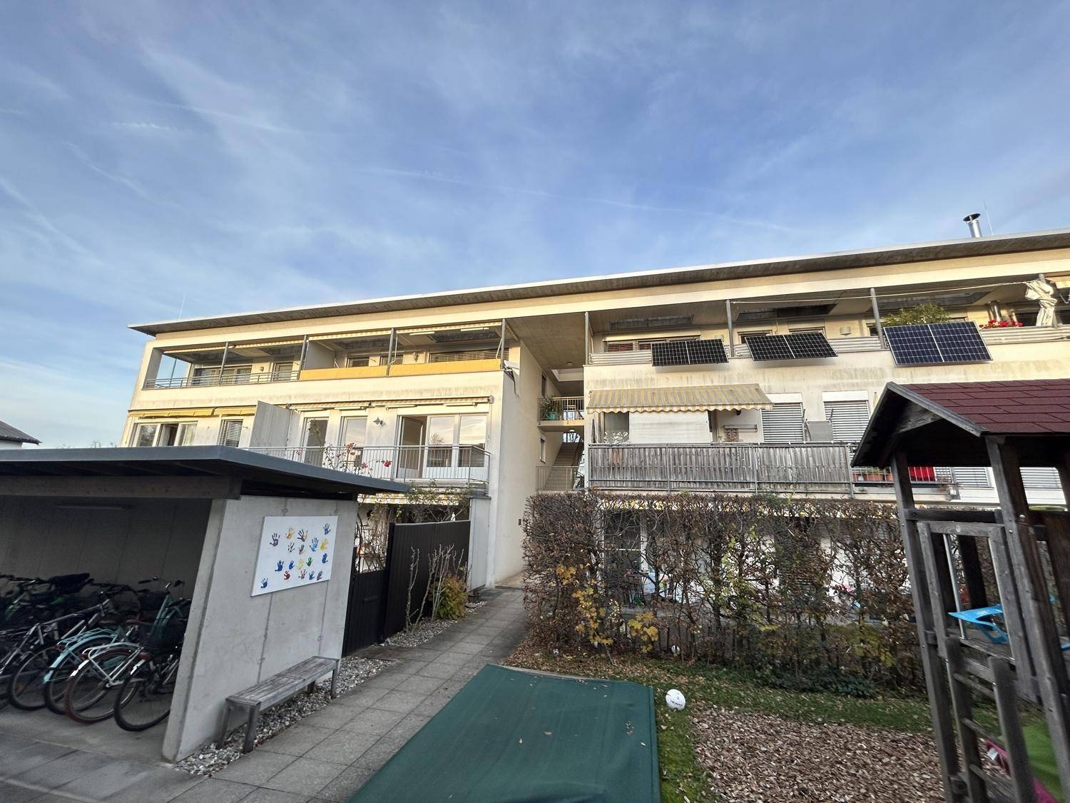 Rückansicht eines Wohngebäudes mit Balkonen, Solaranlagen, Fahrradunterstand und Spielplatz.