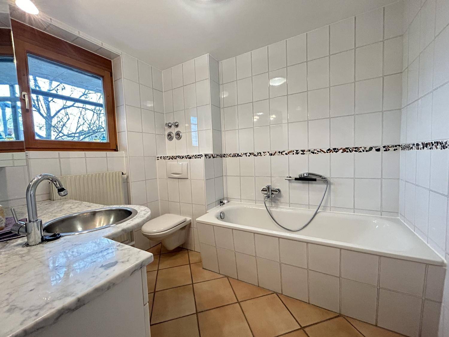 Gefliestes Badezimmer mit Badewanne, Waschtisch mit Marmorplatte und Fenster für Tageslicht.