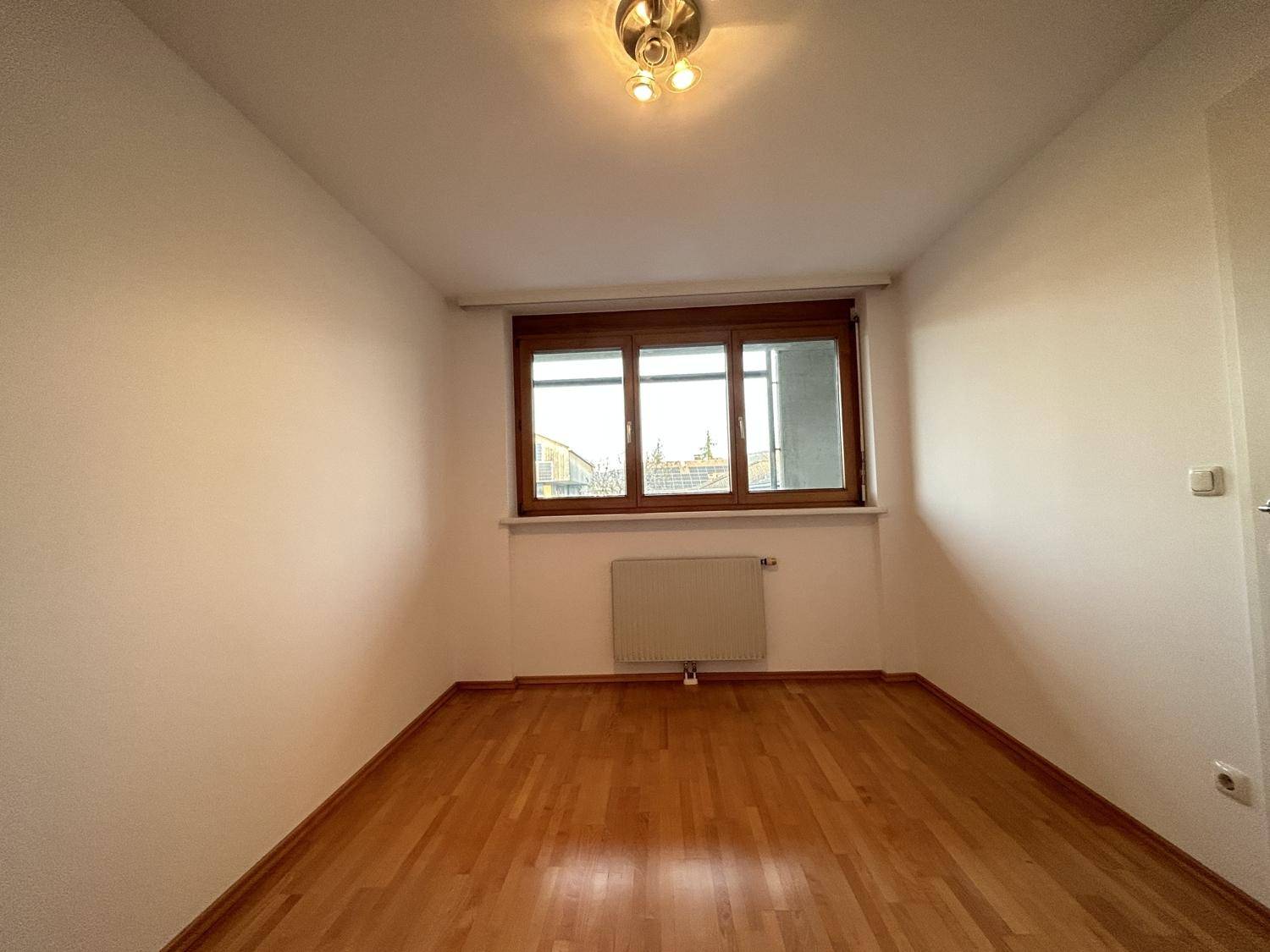 Helles Zimmer mit Holzparkettboden und einem Fenster, das viel Tageslicht hereinlässt.