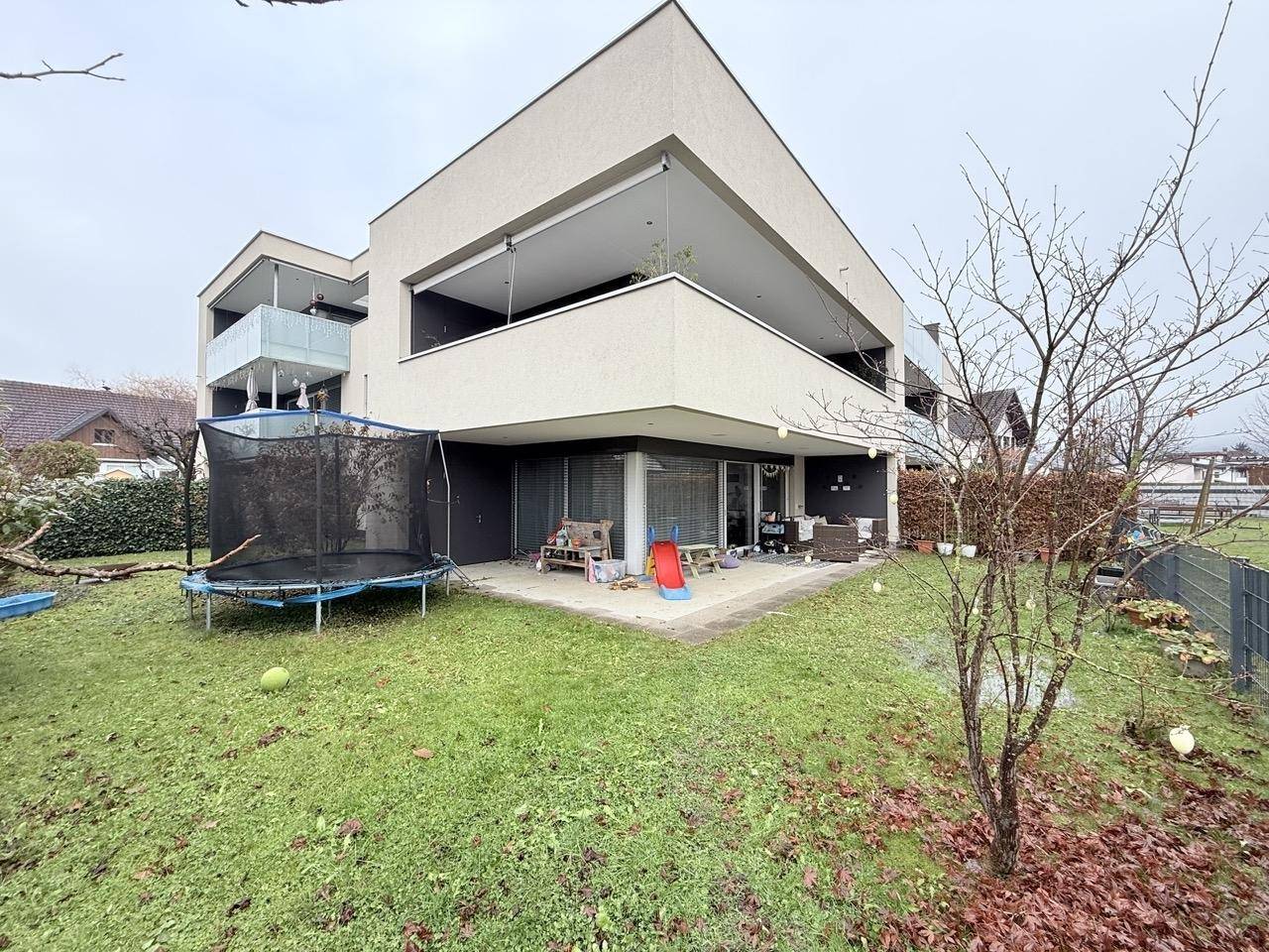 Großer Garten mit Rasenfläche, Trampolin und Terrasse am modernen Gebäude.