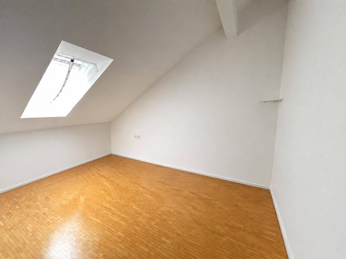 Helles Dachzimmer mit schräger Decke, Dachfenster und Holzboden, ideal als Schlafzimmer.