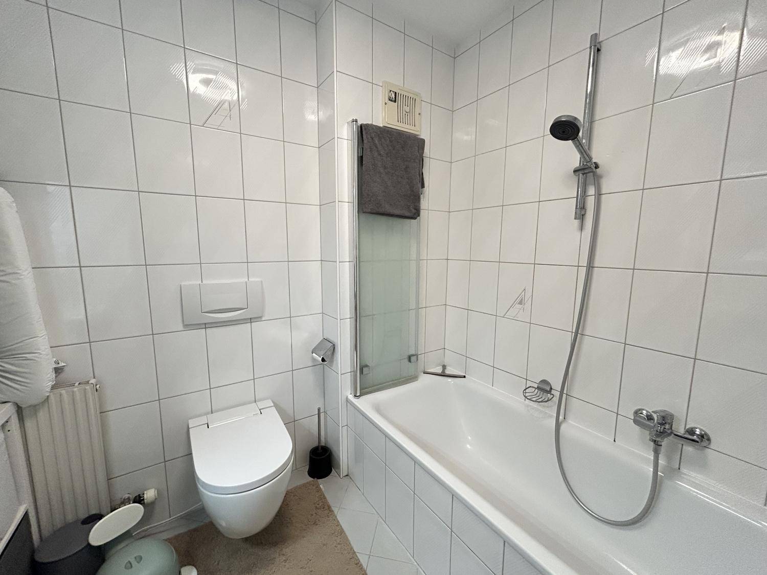 Funktionales Badezimmer mit weißen Fliesen, Badewanne mit Duschfunktion und WC.
