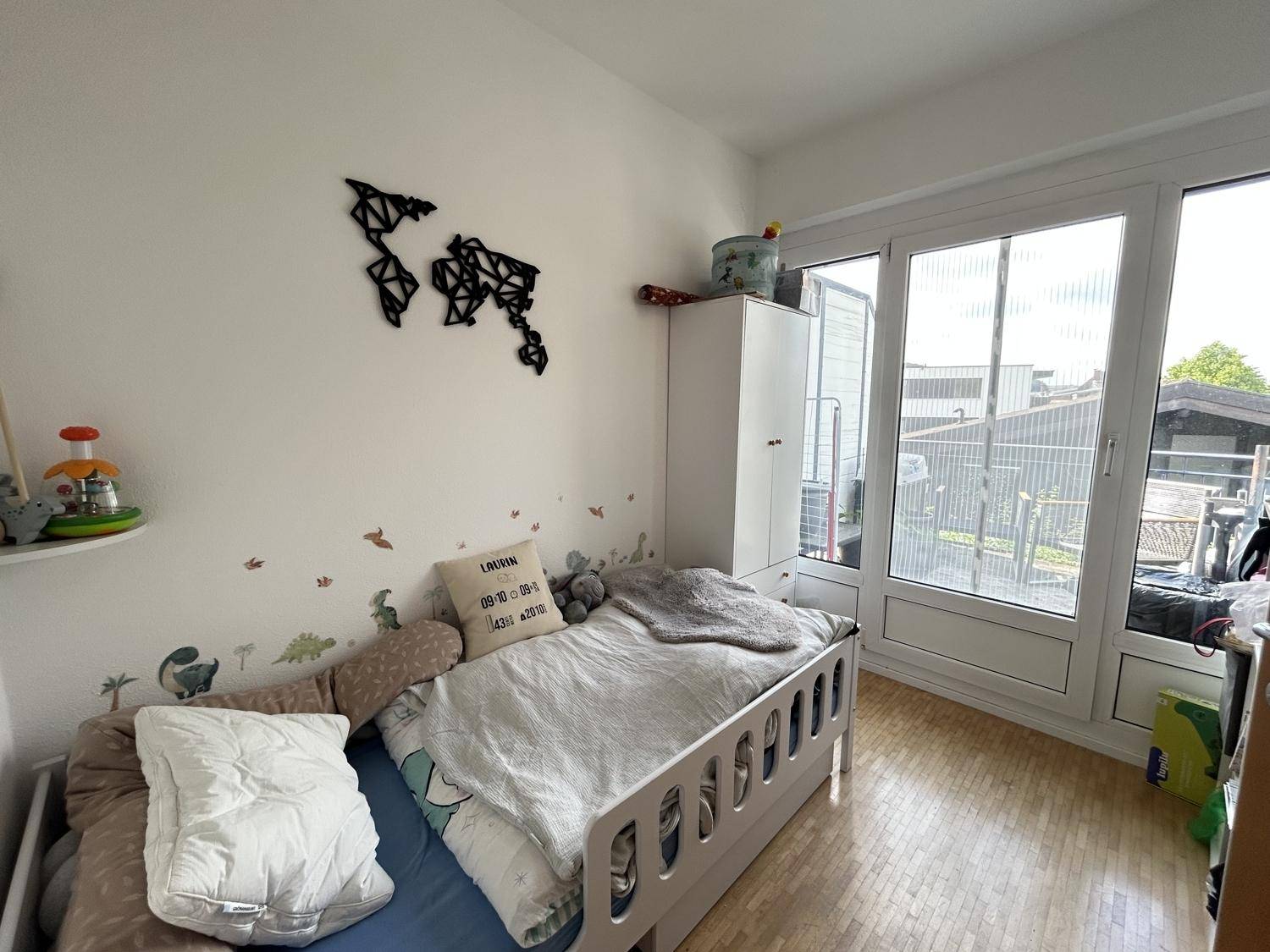 Kinderzimmer mit Bett, Kleiderschrank, großen Fenstern und verspielter Wanddekoration.