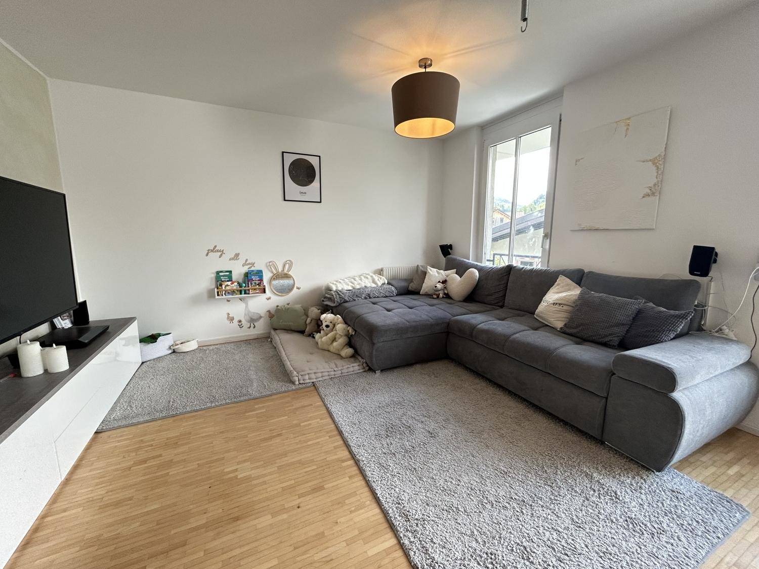 Gemütliches Wohnzimmer mit großem Ecksofa, Holzboden und viel Tageslicht durch die Fenster.