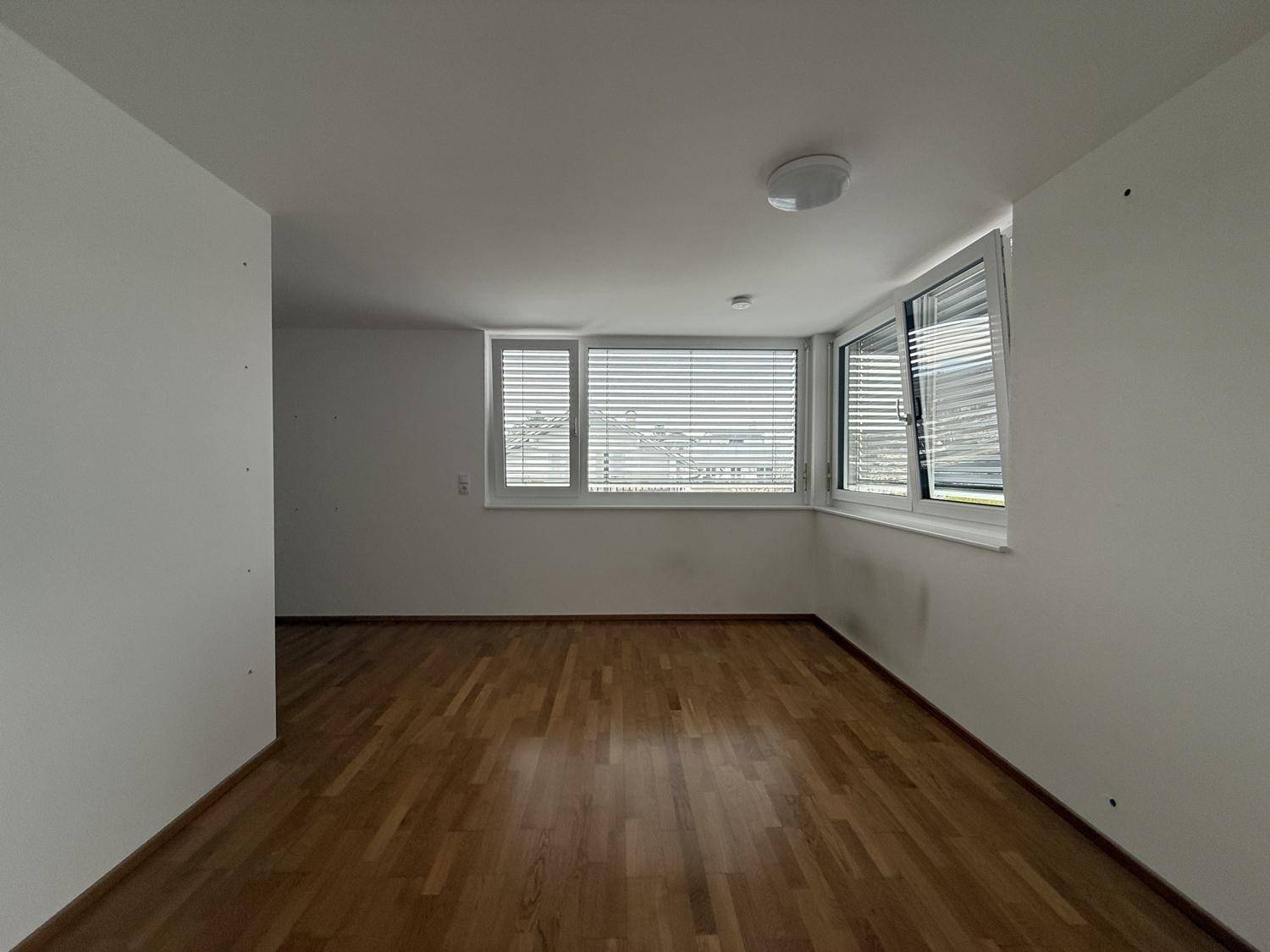 Helles Zimmer mit Holzparkett und breiter Fensterfront, ausgestattet mit Jalousien für Privatsphäre.