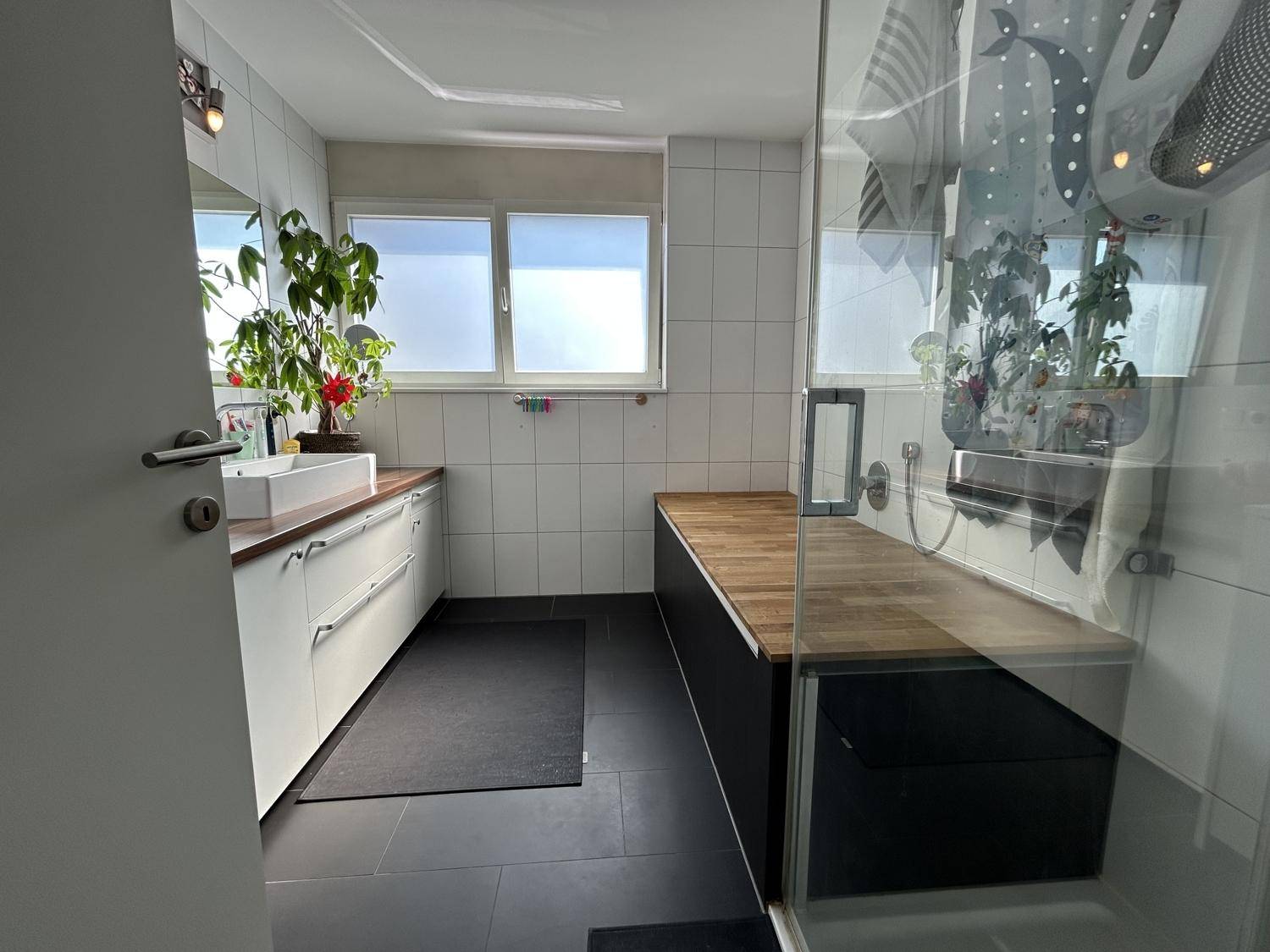 Helles Badezimmer mit großer Dusche, Fenster und modernem Waschtisch mit Holzplatte.