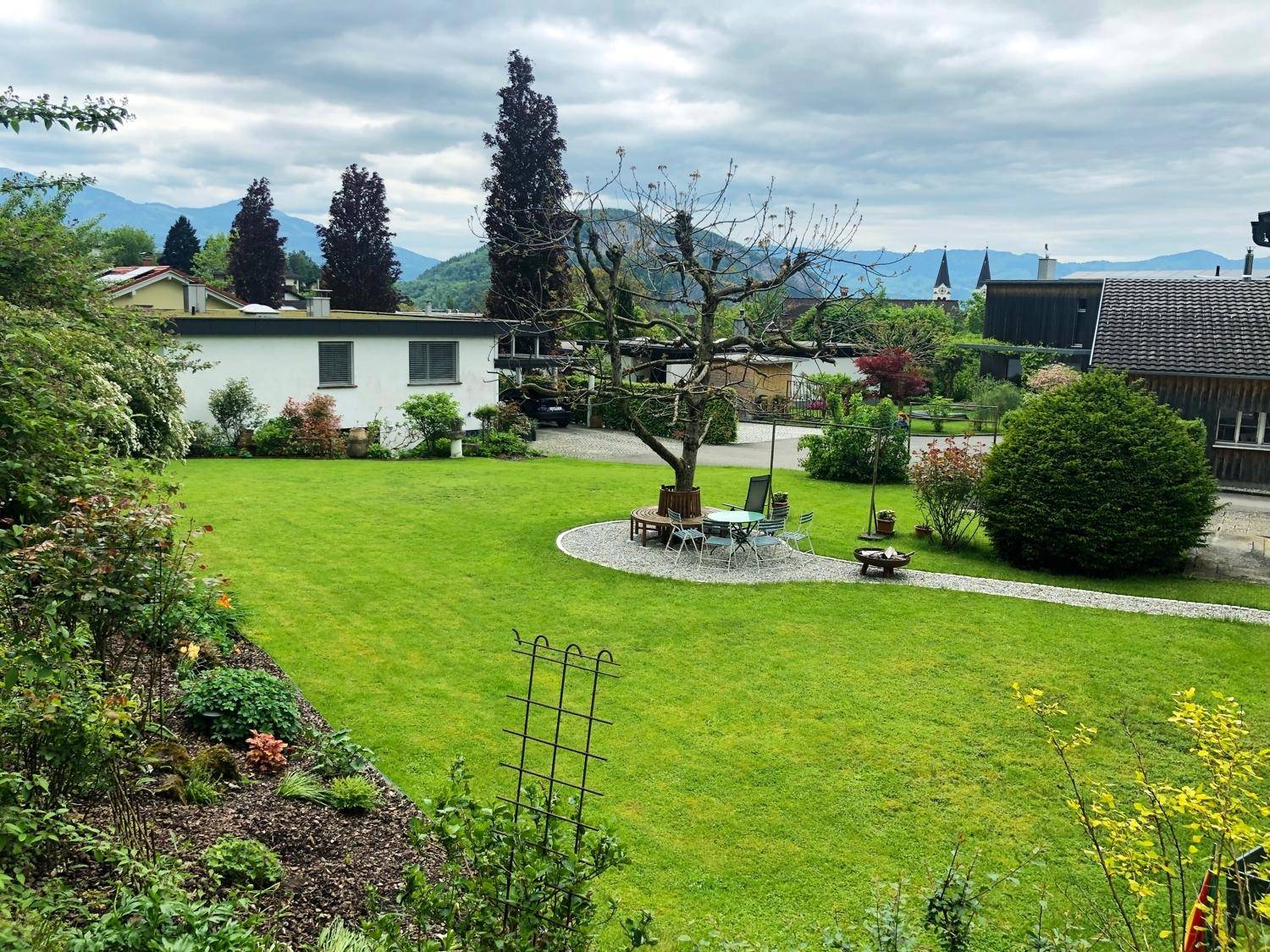 Weitläufiger Garten mit Sitzbereich, Rasenfläche und Fernblick auf Berge und umliegende Häuser.