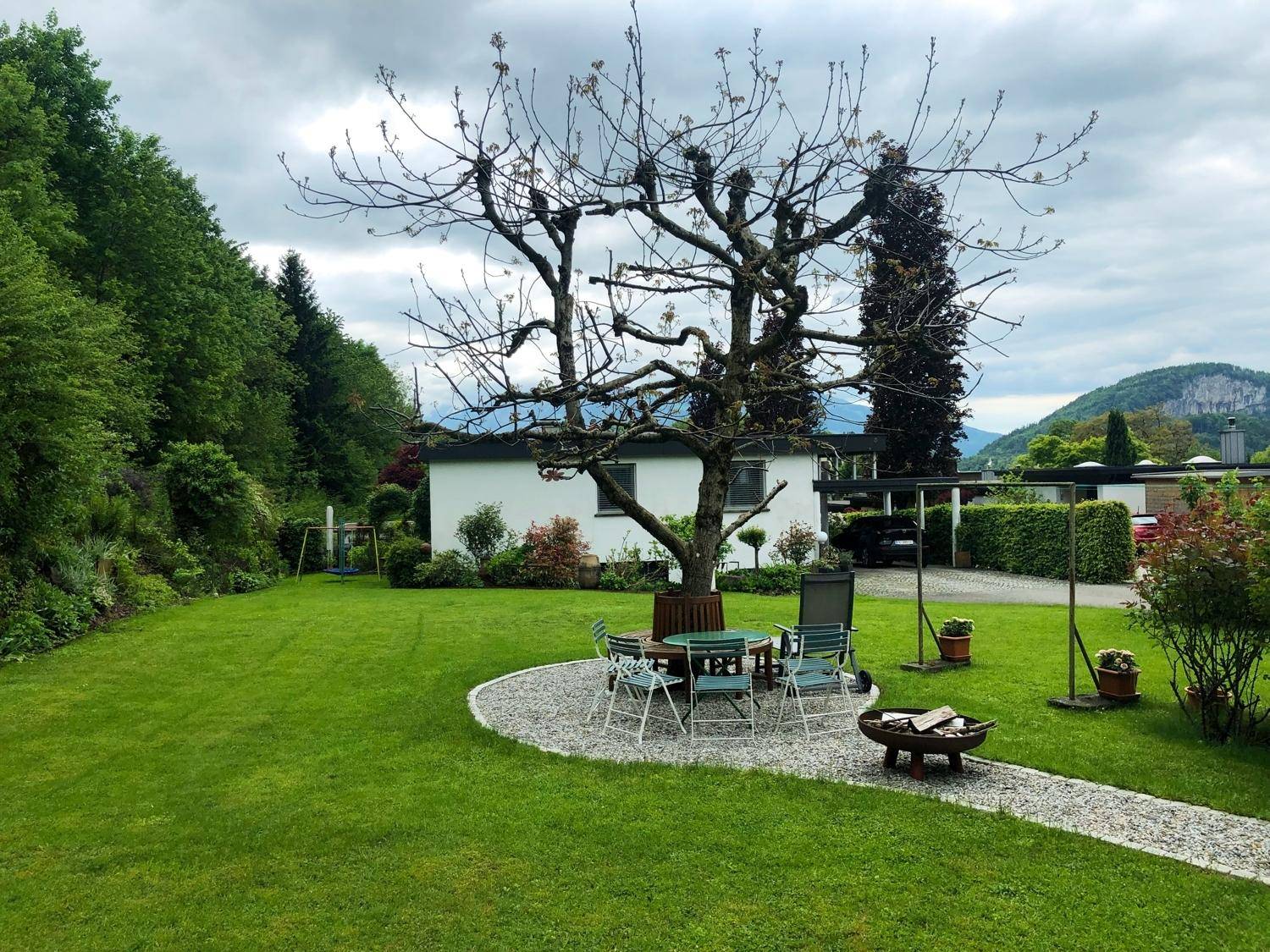 Großer Garten mit Rasenfläche, gemütlichem Sitzbereich und Feuerstelle unter einem Baum.