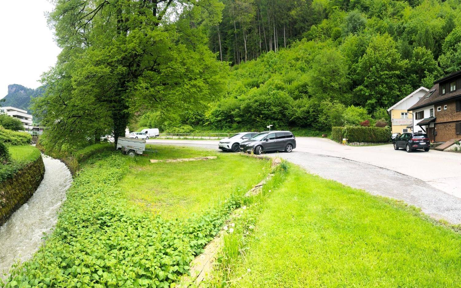 Kleine Straße mit parkenden Autos, einem Bachlauf und einem bewaldeten Berghang im Hintergrund.