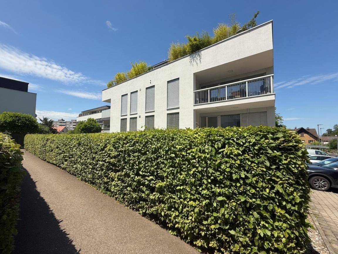 Modernes Wohnhaus mit Balkonen und großen Fenstern, umgeben von einer dichten grünen Hecke.