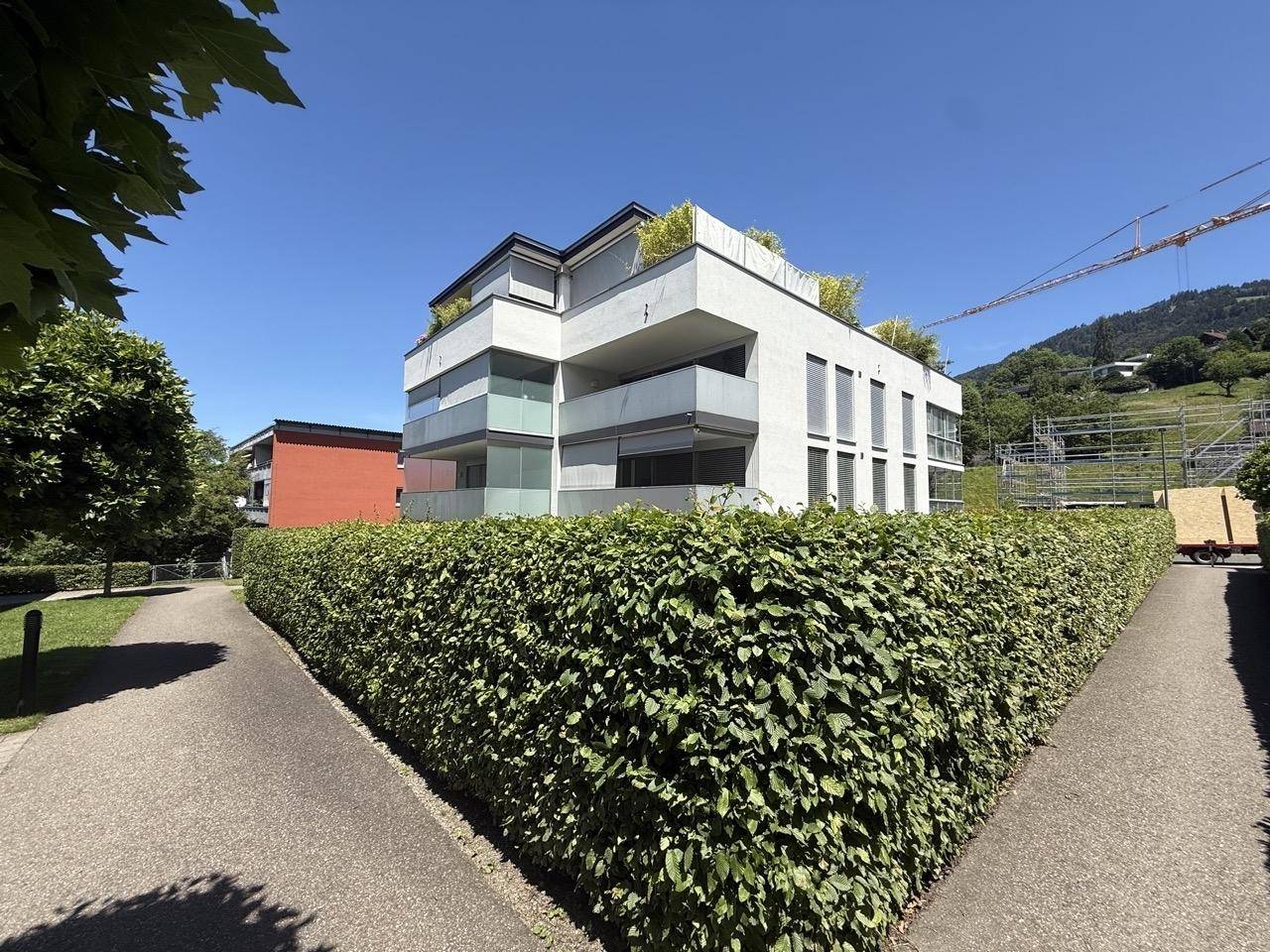 Zeitgenössisches Gebäude mit weißer Fassade und Balkonen, umgeben von üppigem Grün und Hecken.