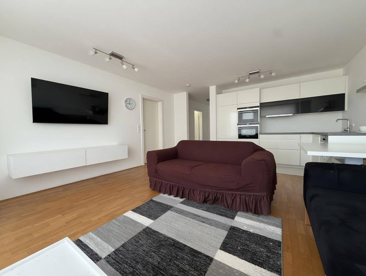Offener Wohnbereich mit Holzboden, Sofa und integrierter moderner Küche im Hintergrund, hell und freundlich.