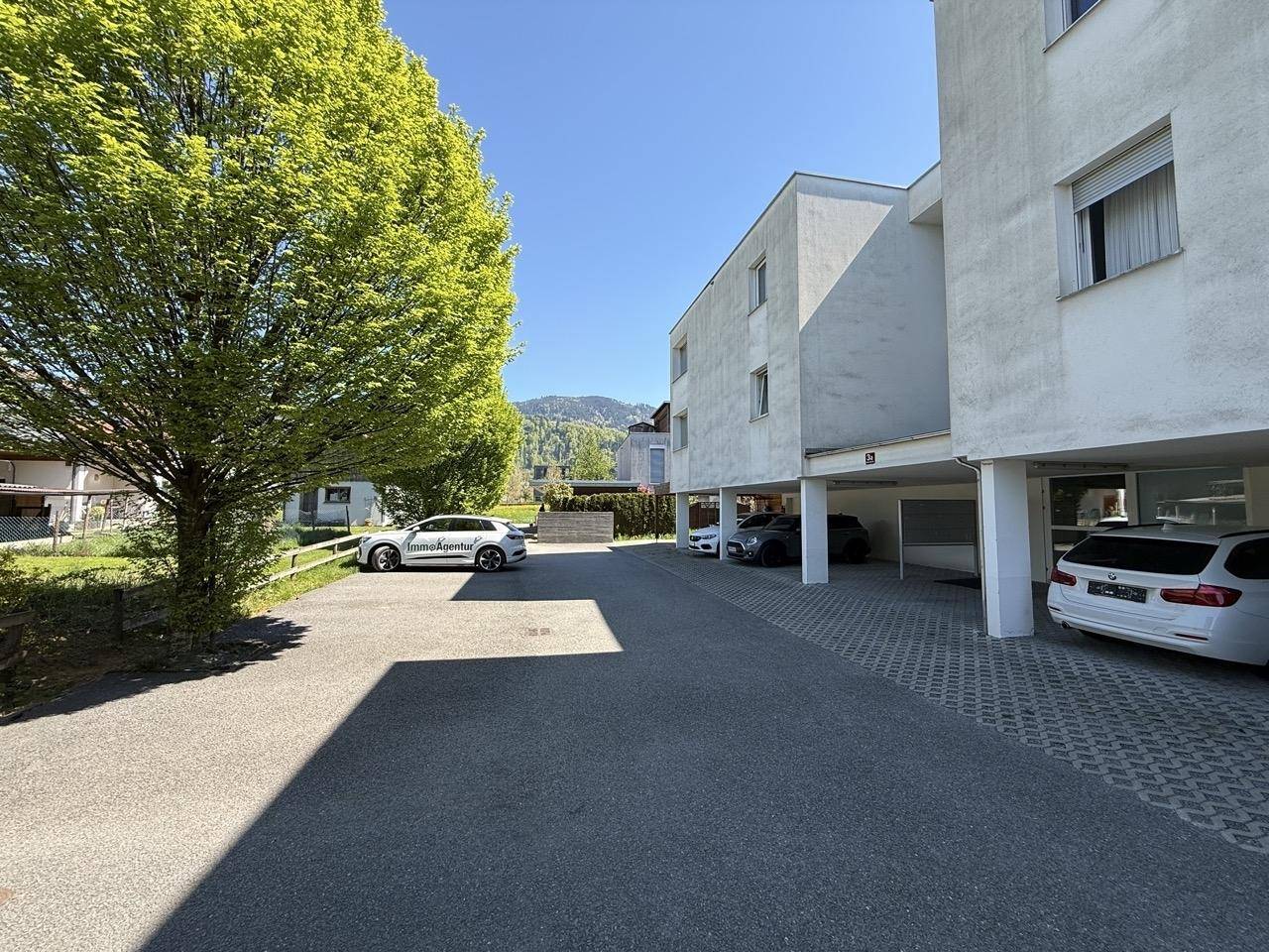 Modernes Mehrfamilienhaus mit Außenparkplätzen und Carports, umgeben von grüner Landschaft und Bergen.