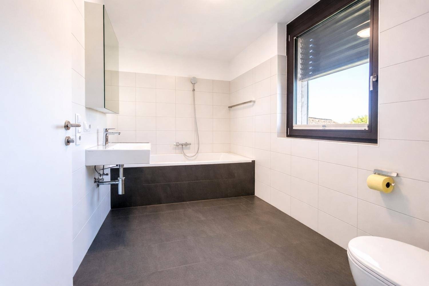 Helles Badezimmer mit Badewanne, WC, Waschbecken und Fenster, ausgestattet mit Fliesen.