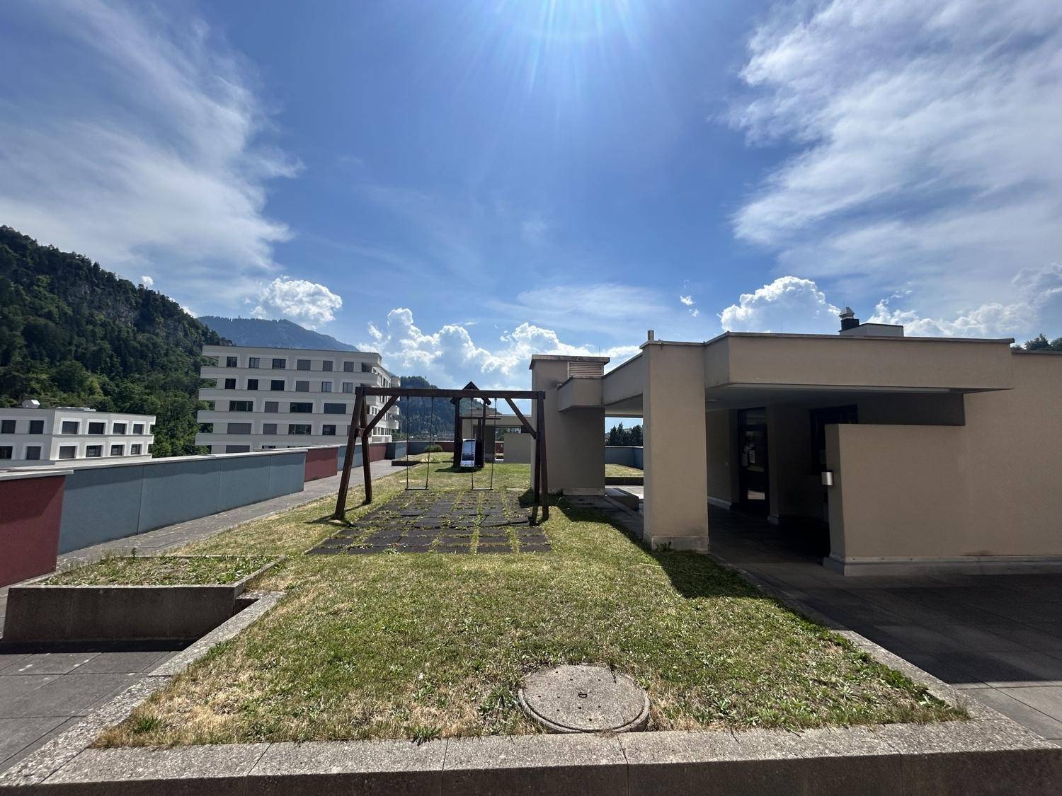 Dachterrasse mit Spielplatz und Grünflächen, umgeben von Gebäuden und Bergen.
