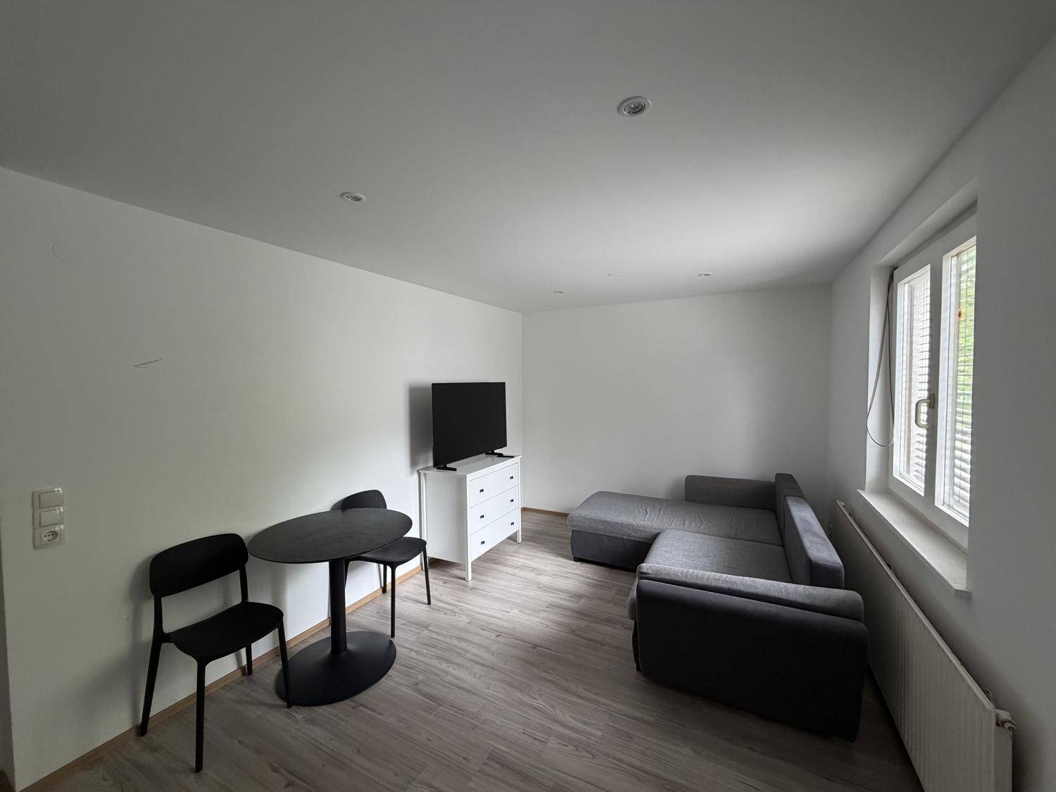 Schlichter Wohnraum mit grauem Sofa, Fernseher und einem kleinen Essbereich am Fenster.