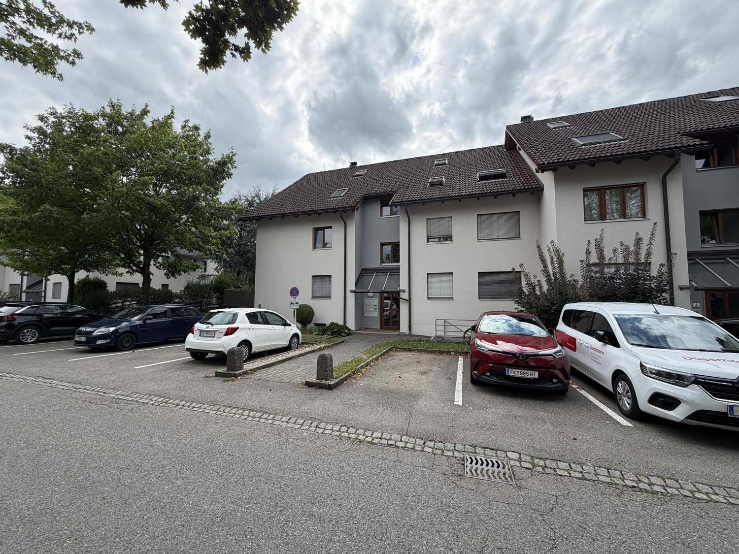 Mehrfamilienhaus mit Parkplätzen vor dem Gebäude unter bewölktem Himmel.