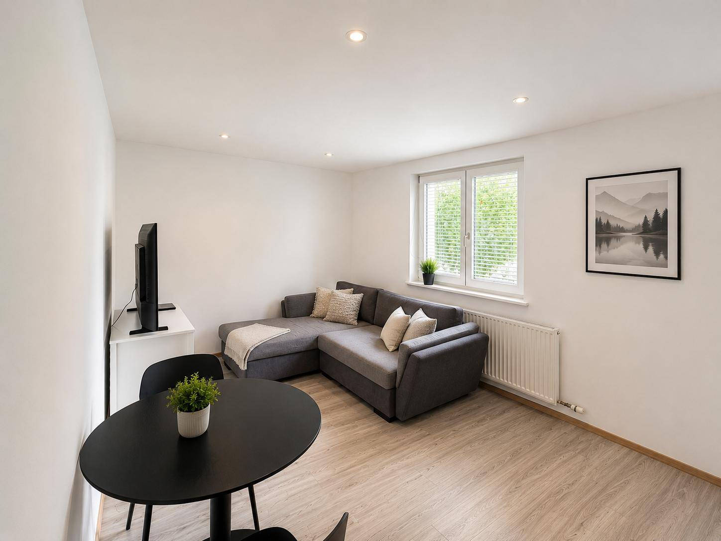 Gemütliches Wohnzimmer mit grauem Ecksofa, kleinem Esstisch und hellem Laminatboden.