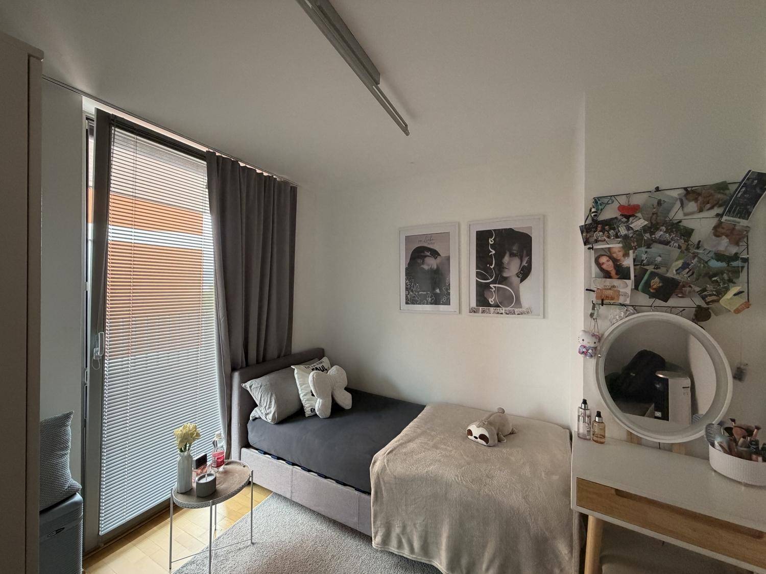 Helles Schlafzimmer mit Einzelbett, Holzboden und großem Fenster mit Jalousien sowie einem Schminktisch.