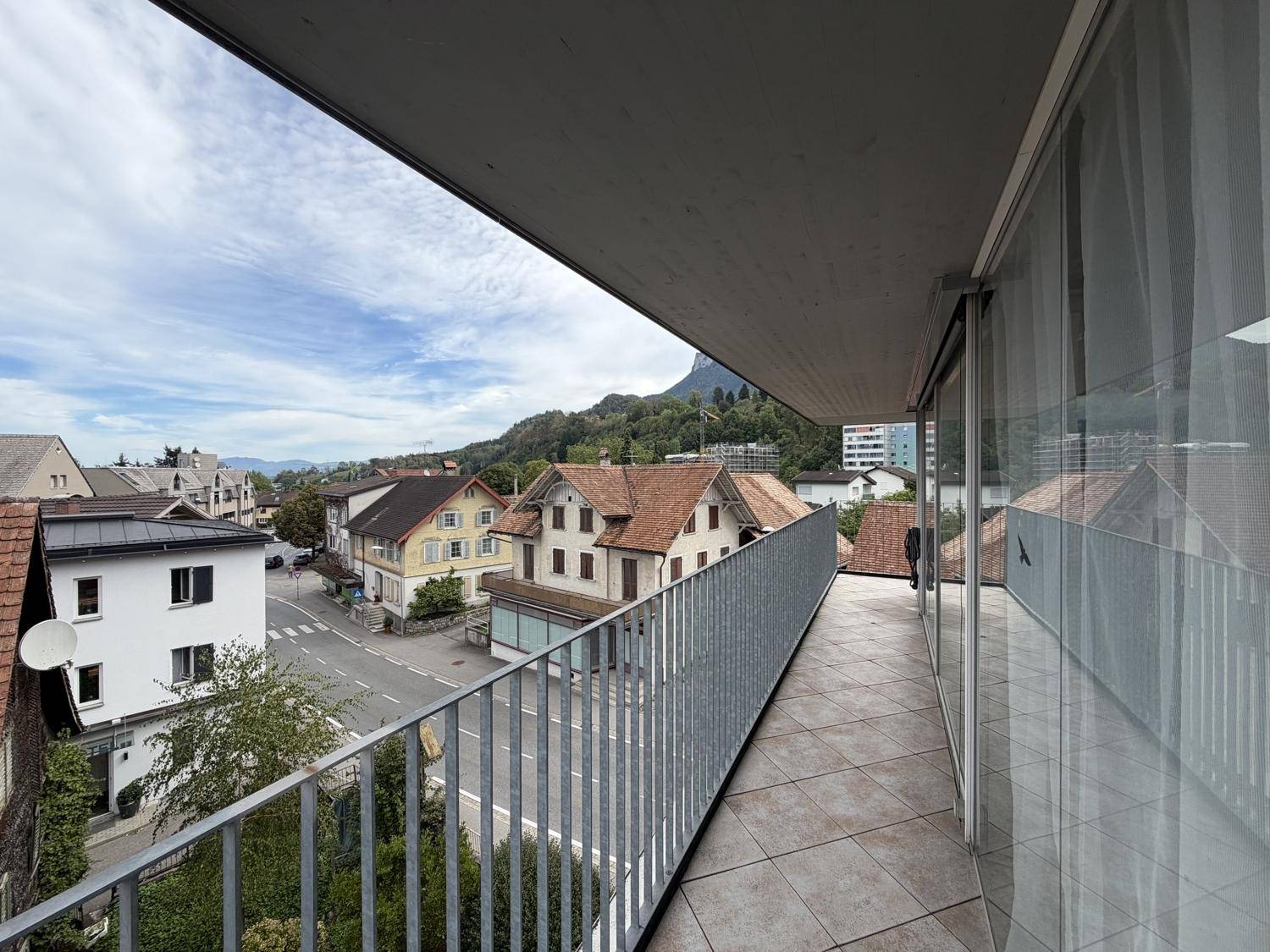 Langer Balkon mit Fliesenboden und Geländer, der einen Blick auf die Straße und die umliegenden Häuser bietet.