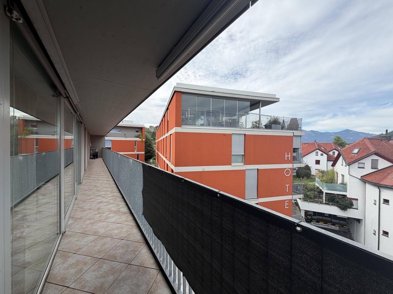 Langer Balkon mit Fliesenboden und Blick auf die umliegenden Gebäude und die Berglandschaft.