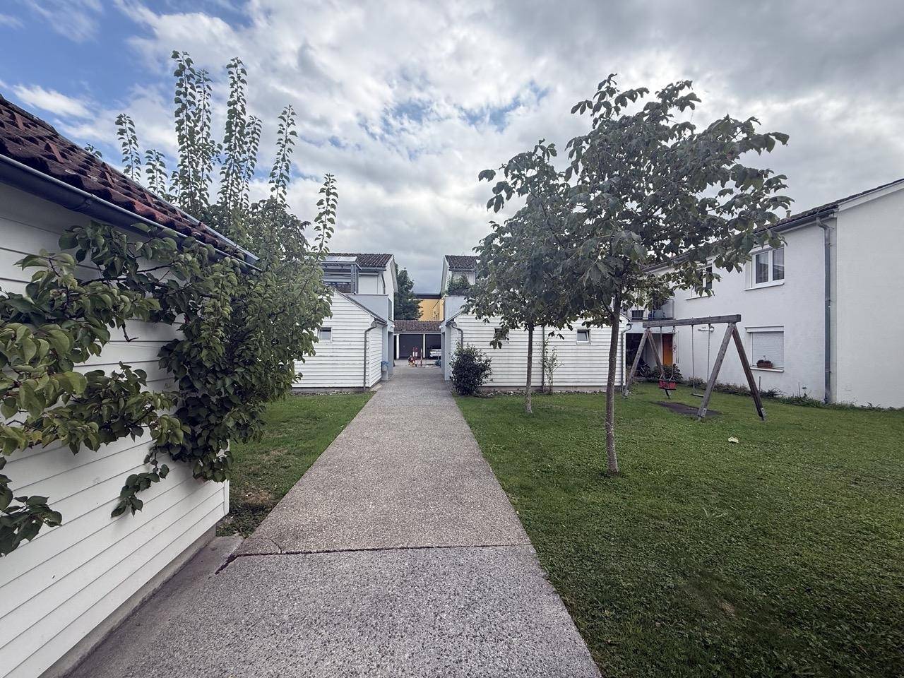 Grüner Gartenbereich mit einem Weg zwischen den Gebäuden und einer Schaukel im Hintergrund.