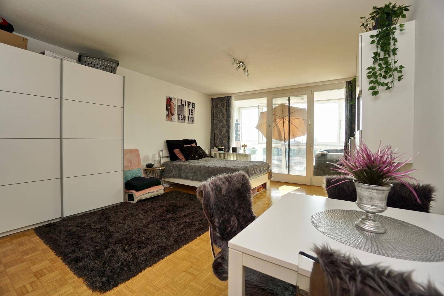 Heller Wohnbereich in einem Studio-Apartment mit Bett, Esstisch und Zugang zum Balkon.