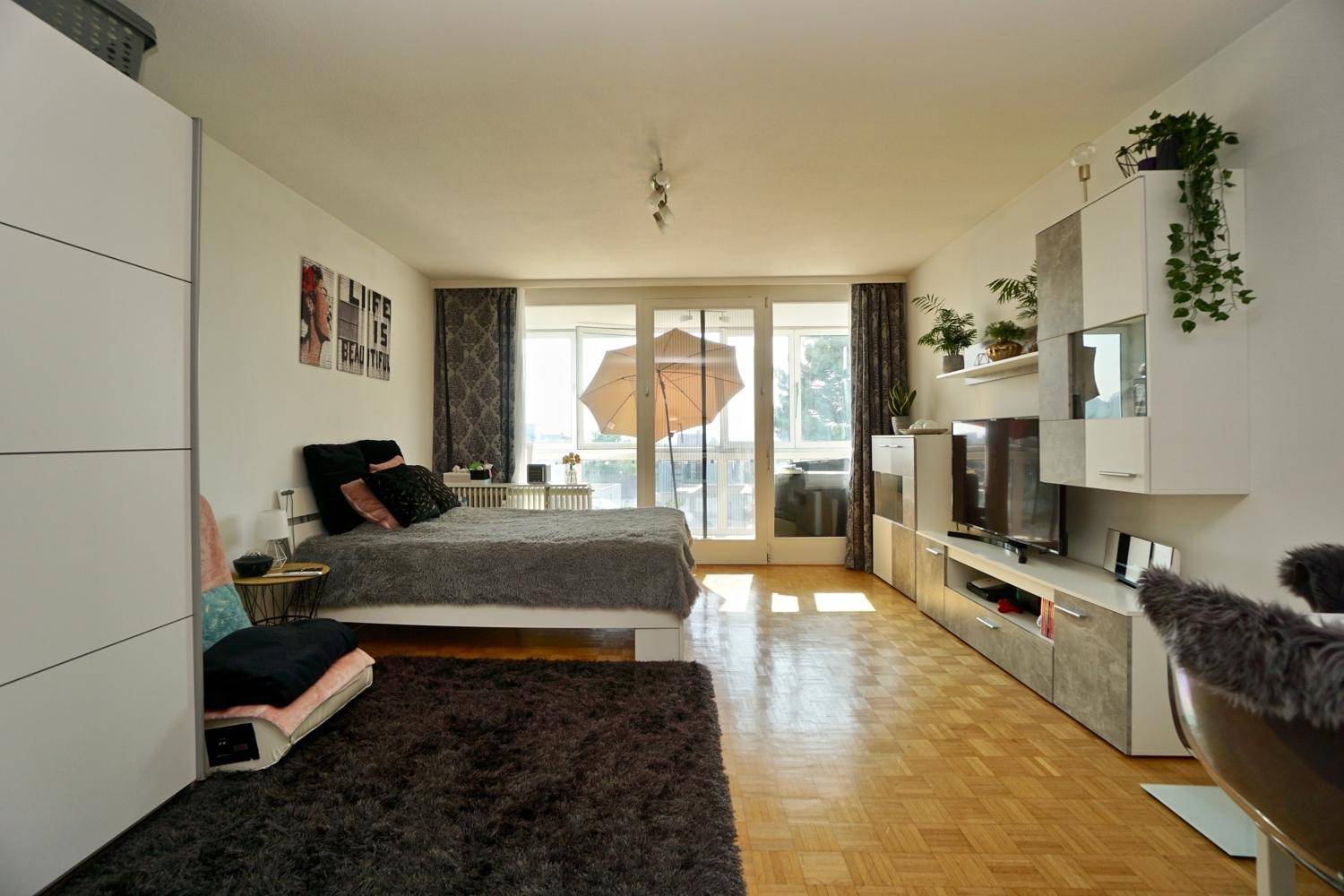 Gemütlicher Wohnbereich mit Bett, TV-Möbeln und großen Fenstern, die viel Tageslicht hereinlassen.