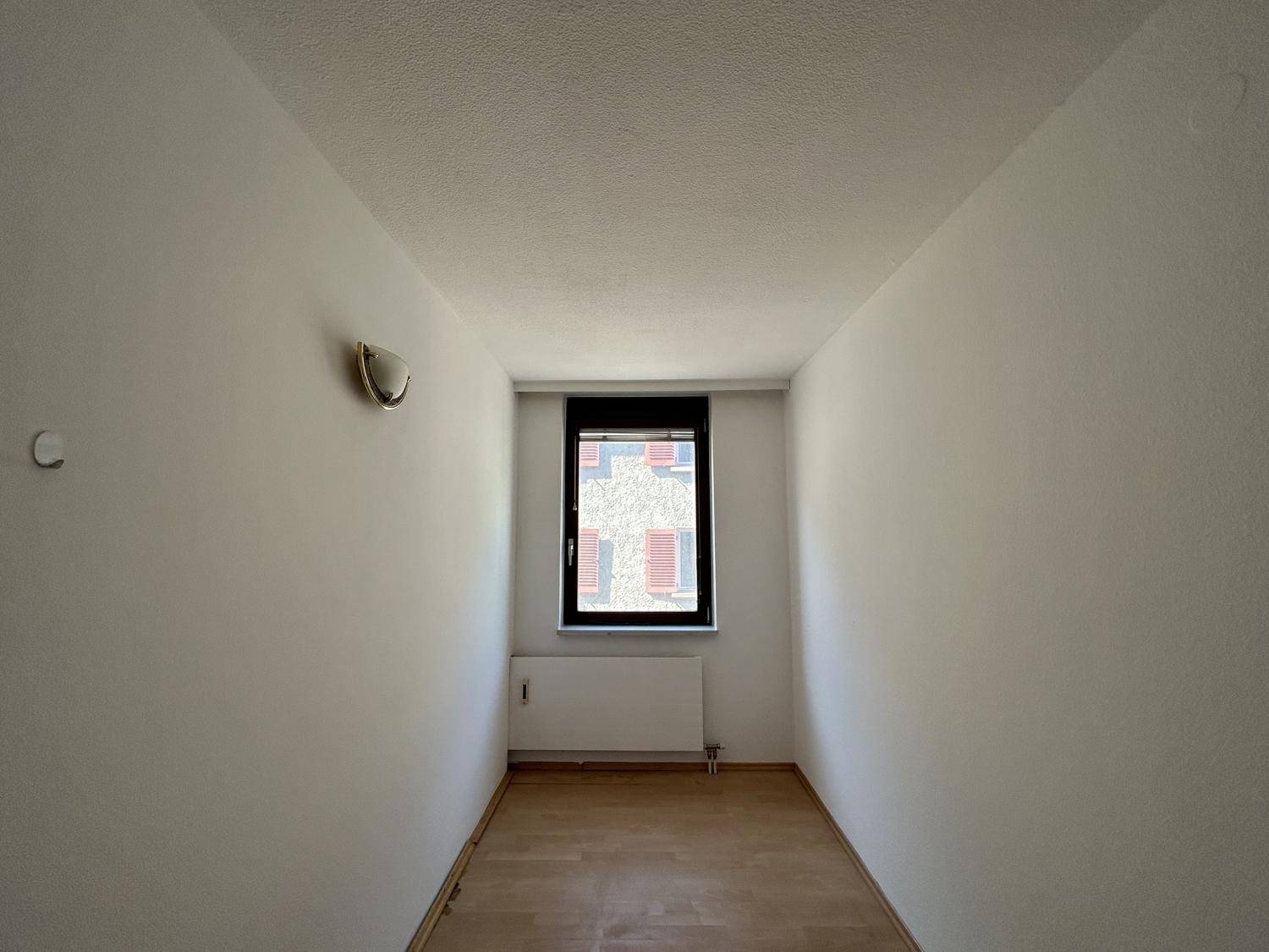 Kleiner, schmaler Raum mit einem Fenster und hellem Laminatboden, ideal als Schlafzimmer.