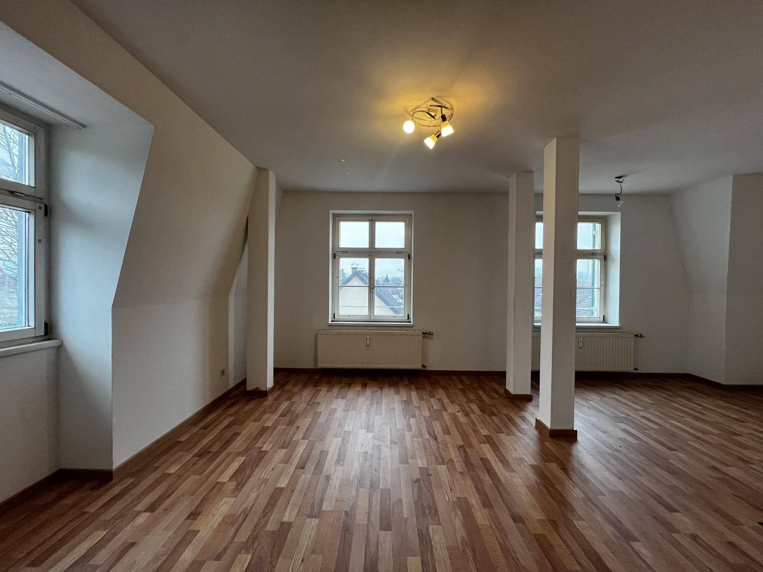 Heller Wohnraum mit Holzboden, hohen Decken und mehreren Fenstern für viel Tageslicht im Dachgeschoss.
