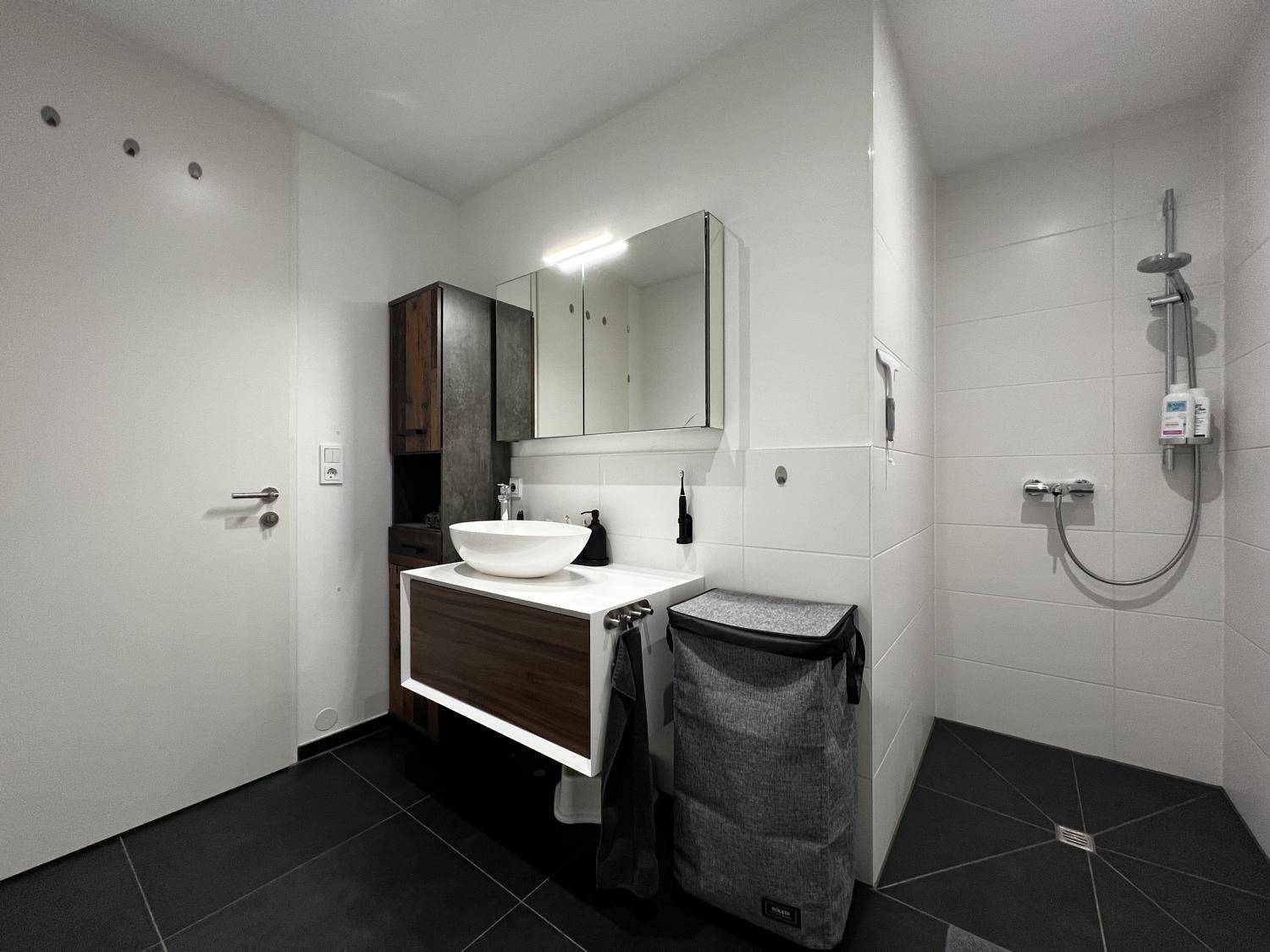 Modernes Badezimmer mit dunklen Fliesen, weißem Waschbecken und ebenerdiger Dusche.