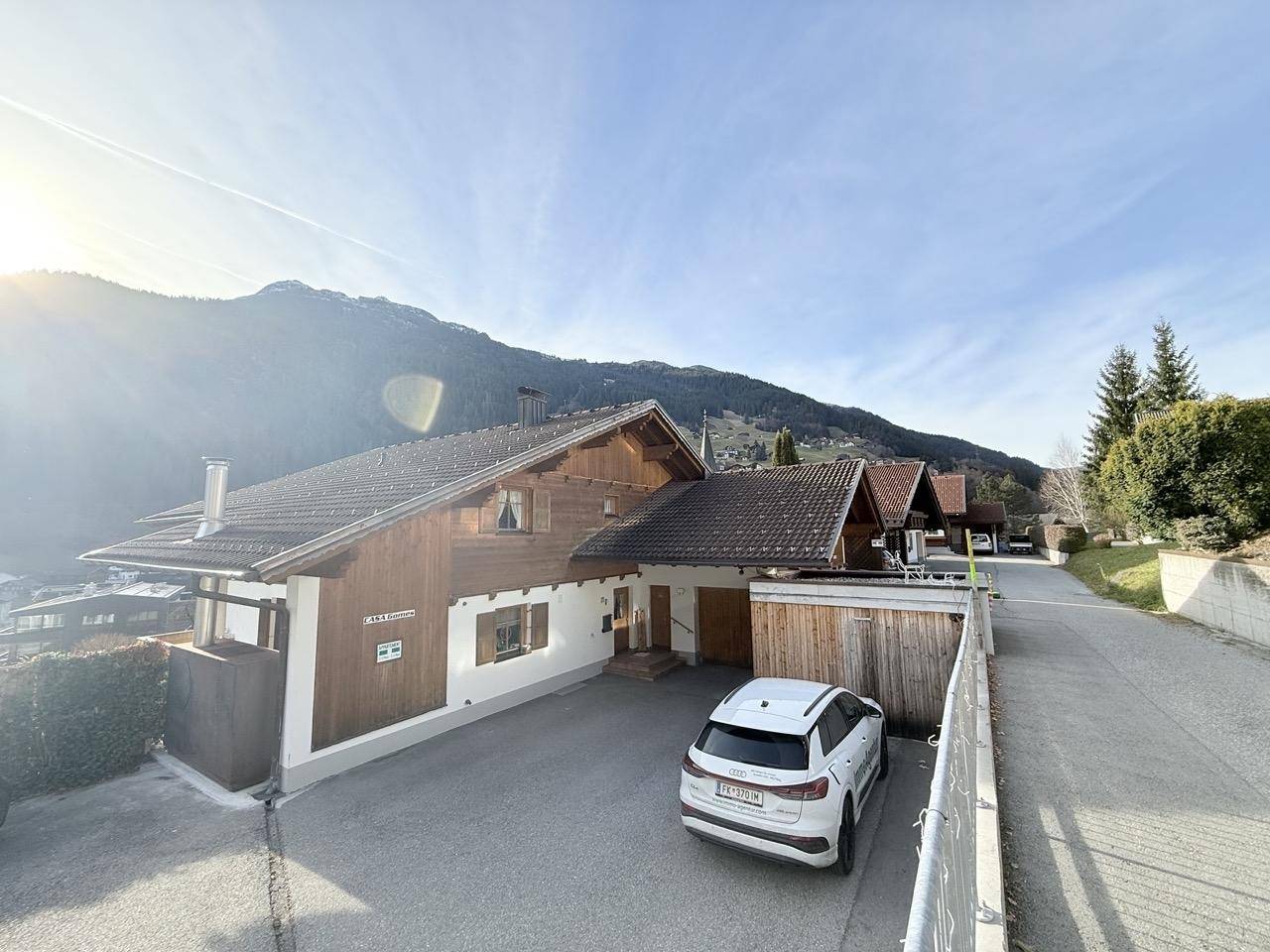 Haus mit breiter Zufahrt und Parkplatz, umgeben von Bergen und sonnigem Himmel.