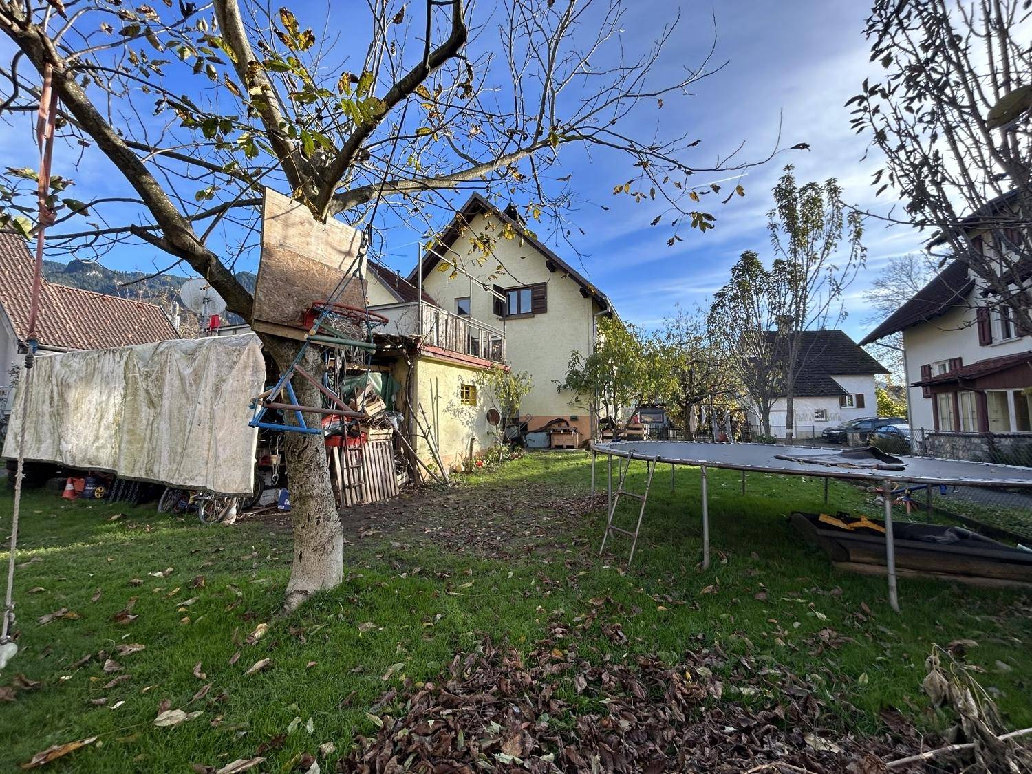 Gartenbereich hinter dem Haus mit Trampolin, einem Baumhaus und herbstlichem Laub auf dem Rasen.