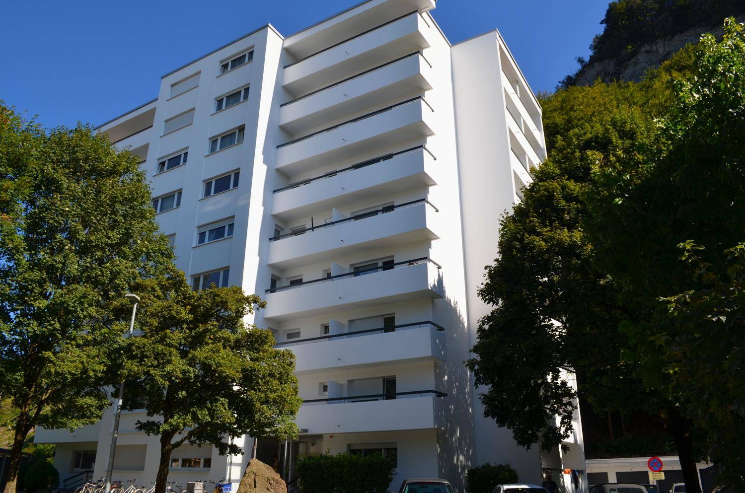 Weißes Mehrfamilienhaus mit vielen Balkonen, umgeben von grünen Bäumen und einem Berg im Hintergrund.