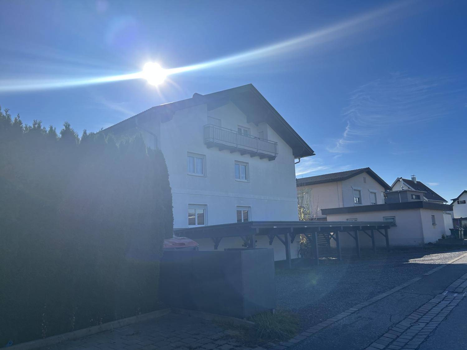 Hausfassade an einem sonnigen Tag mit Carport und Balkon, umgeben von grüner Hecke.