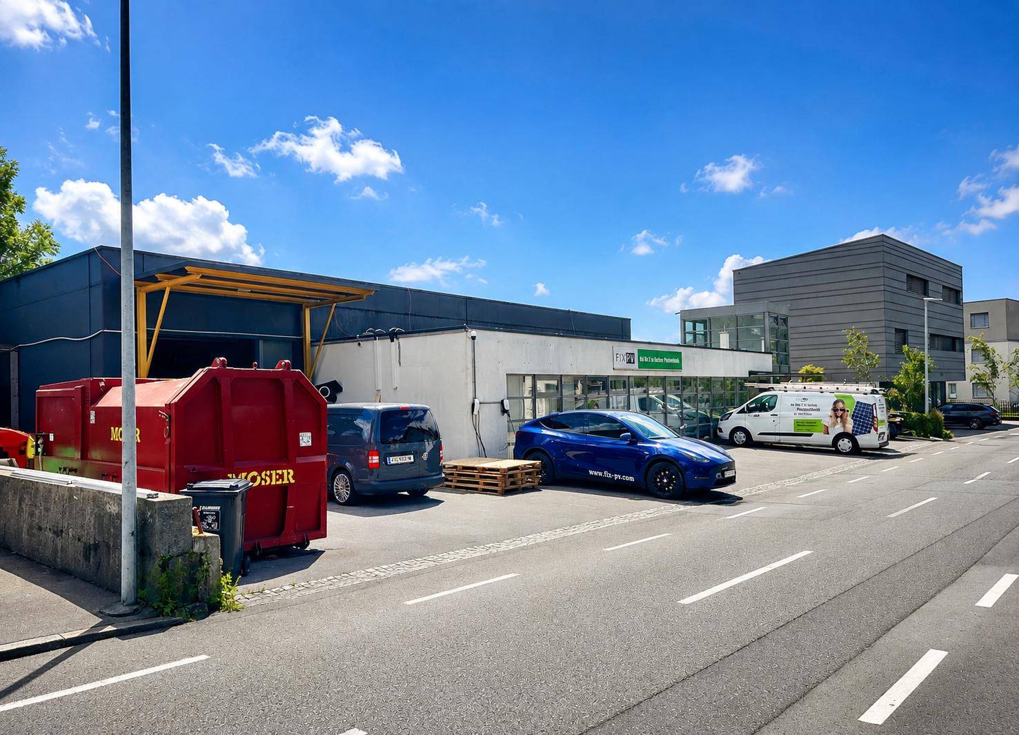 Außenansicht eines modernen Geschäftsgebäudes mit Parkplätzen, einem blauen Elektroauto und einem roten Container.