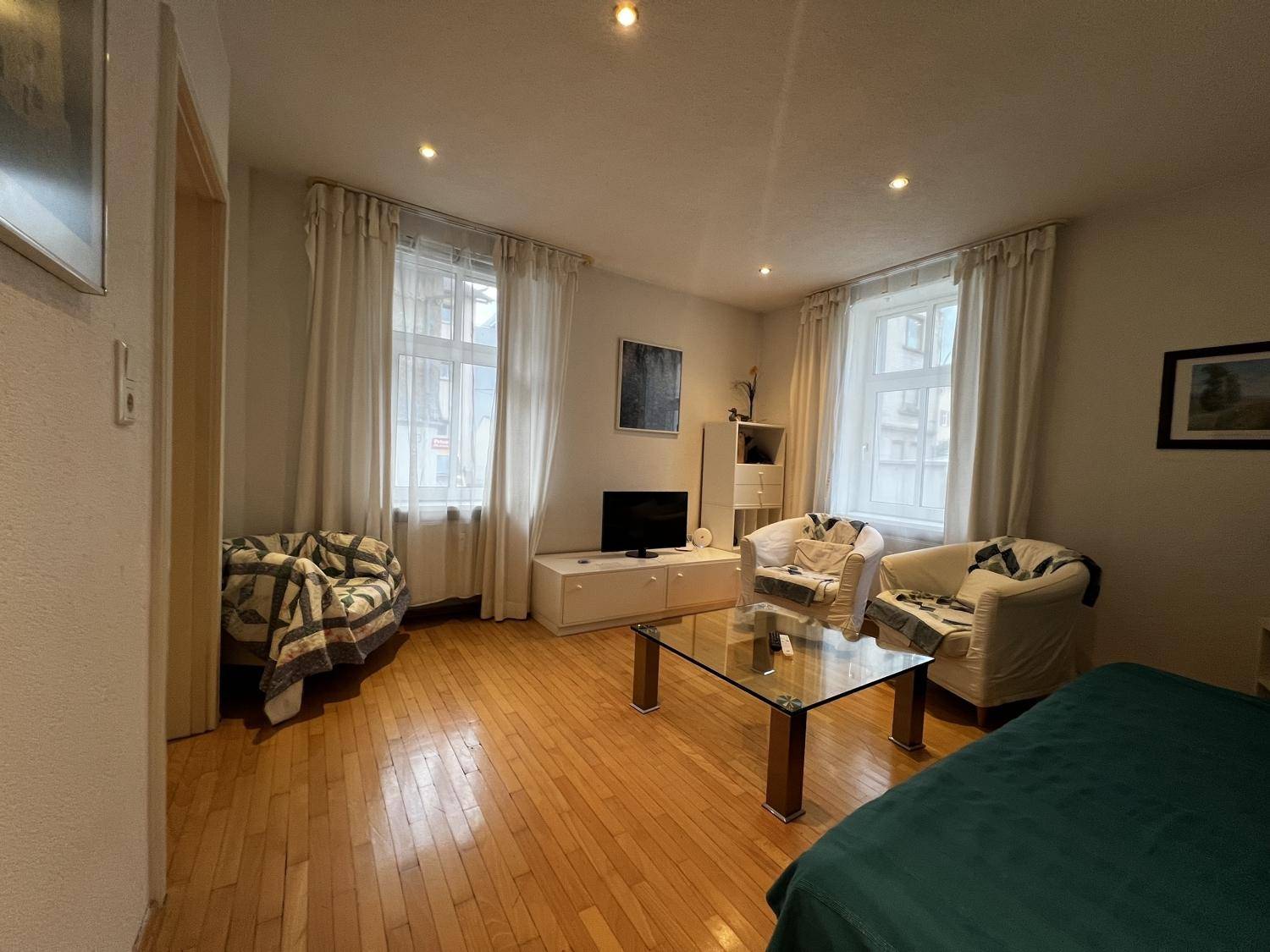 Helles Wohnzimmer mit Holzboden, zwei großen Fenstern und moderner Einrichtung, die zum Verweilen einlädt.