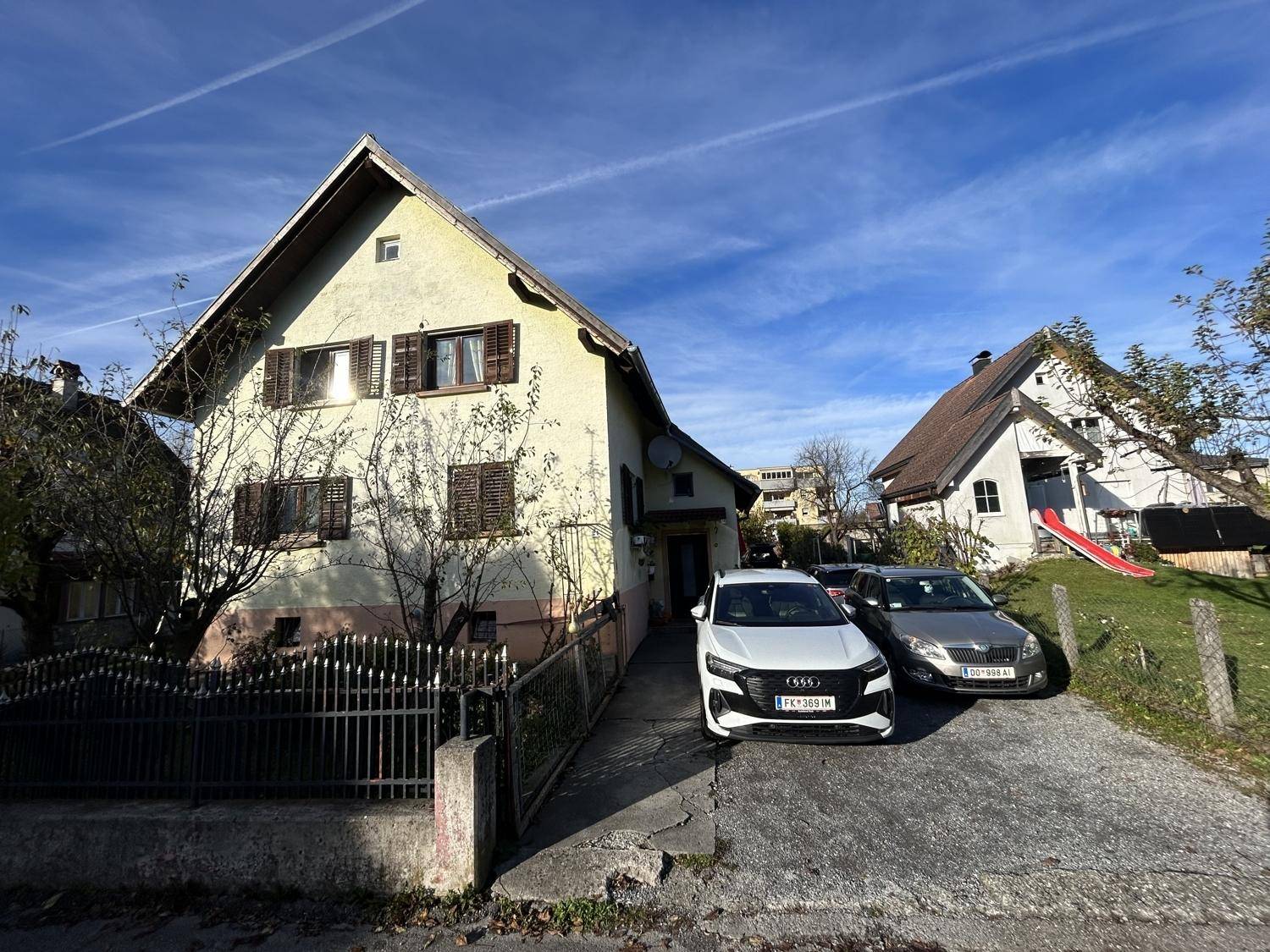 Vorderansicht des Hauses mit Zufahrt, Zaun und geparkten Autos unter klarem blauem Himmel.