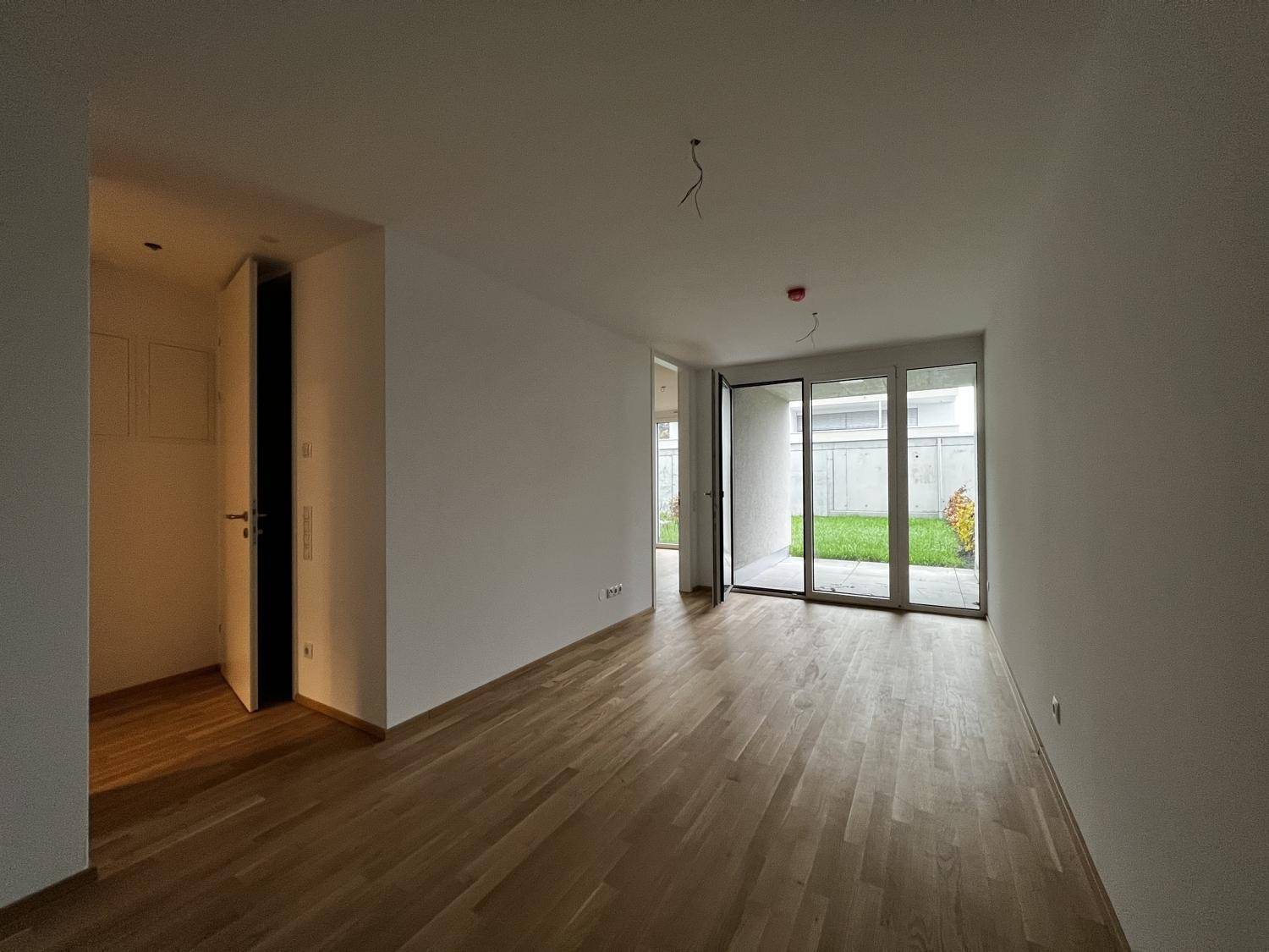 Heller, leerer Raum mit Holzboden und großen Glasschiebetüren zum Gartenbereich.