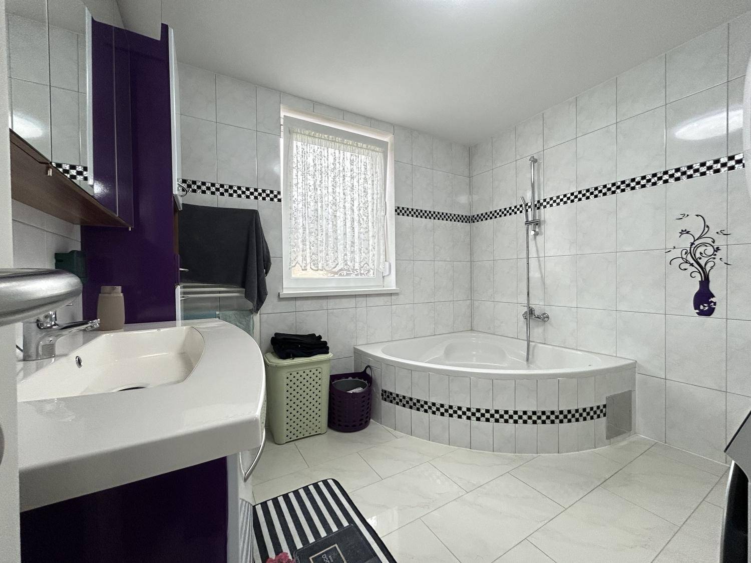 Gefliestes Badezimmer mit Eckbadewanne, Waschbecken und Fenster, dekoriert mit schwarzen Akzenten.