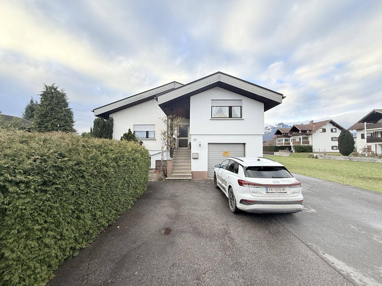 Das Einfamilienhaus mit Einfahrt und Garage, umgeben von grünen Flächen und Bergen im Hintergrund.