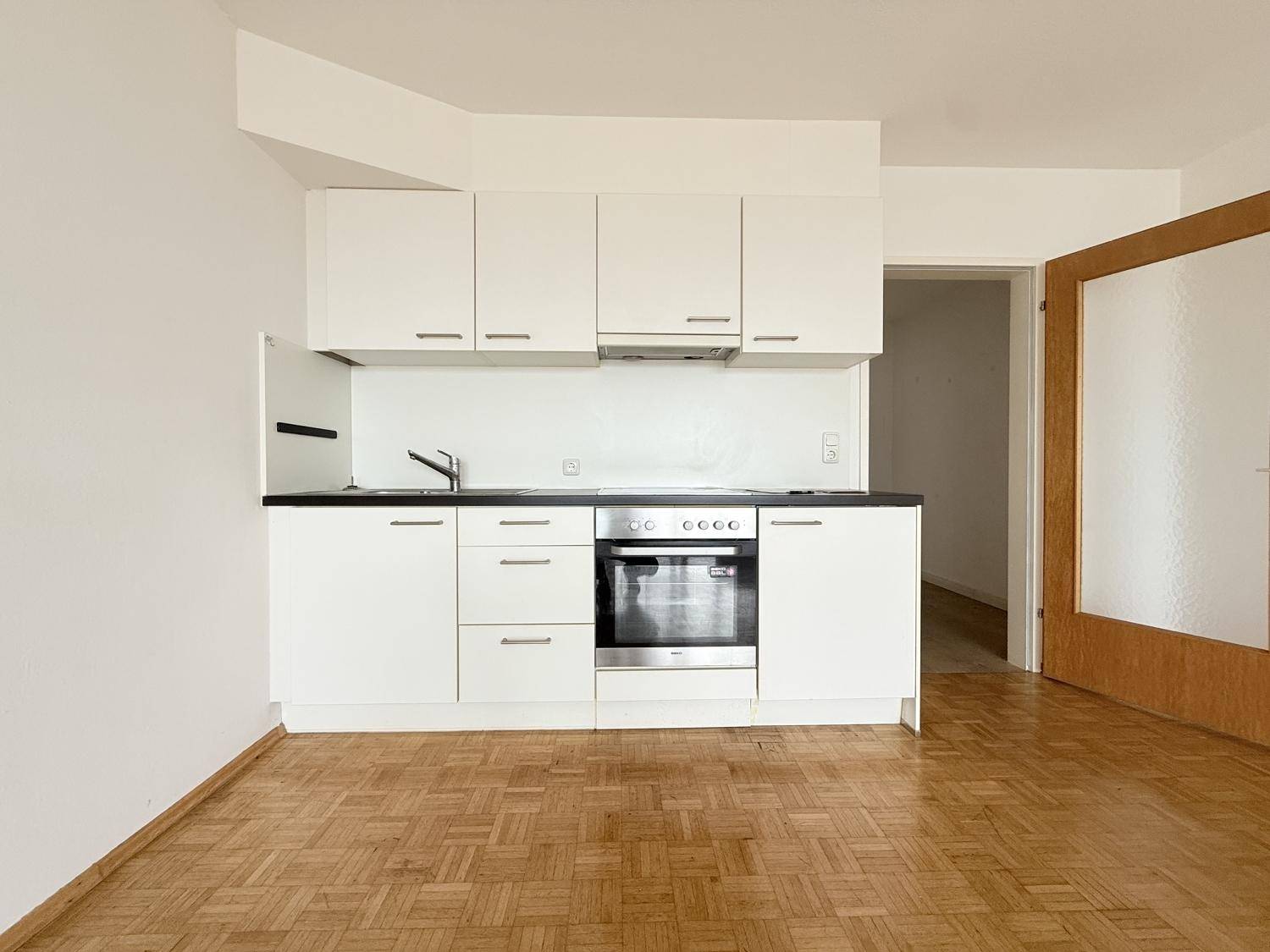 Kompakte Küchenzeile mit weißen Schränken und integriertem Backofen in einem Raum mit Parkettboden.