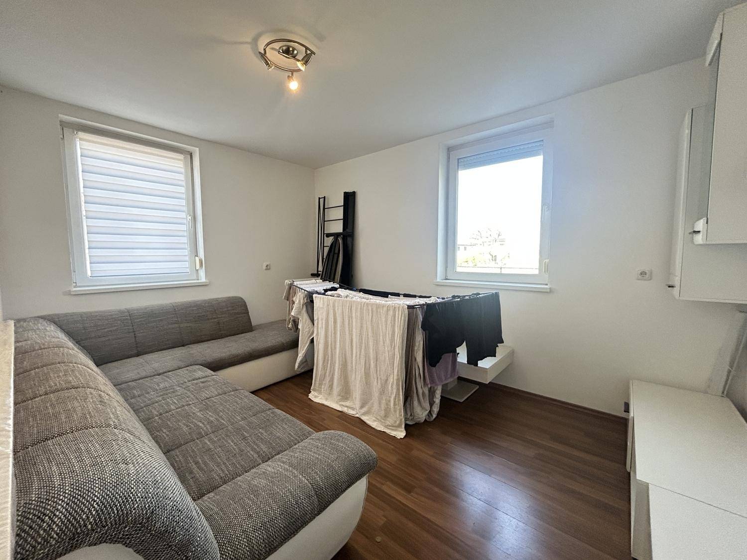 Gemütliches Wohnzimmer mit Ecksofa, zwei Fenstern und Laminatboden, derzeit als Trockenraum genutzt.
