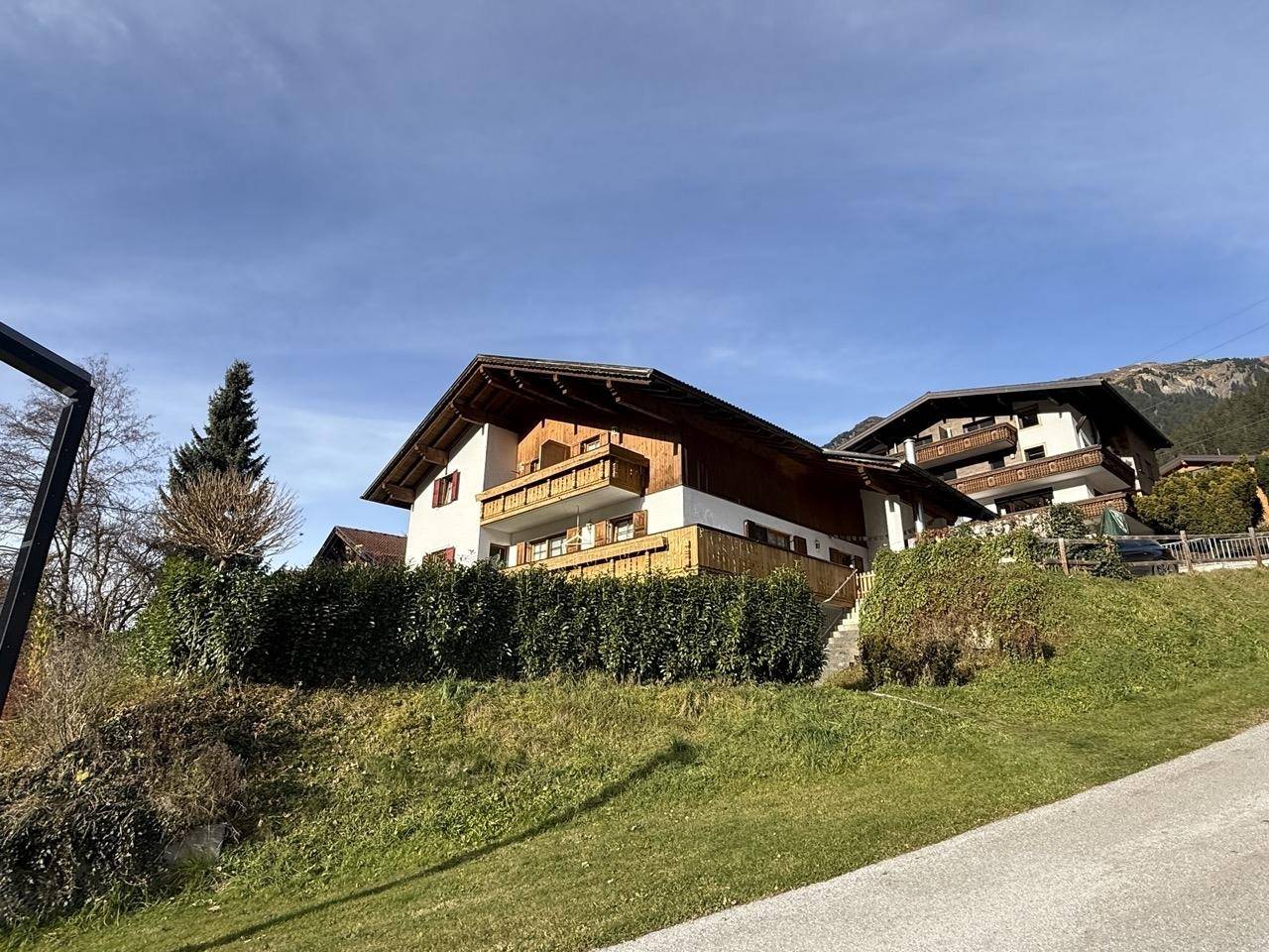 Hausfassade mit Holzelementen und grüner Hecke am Hang unter blauem Himmel.