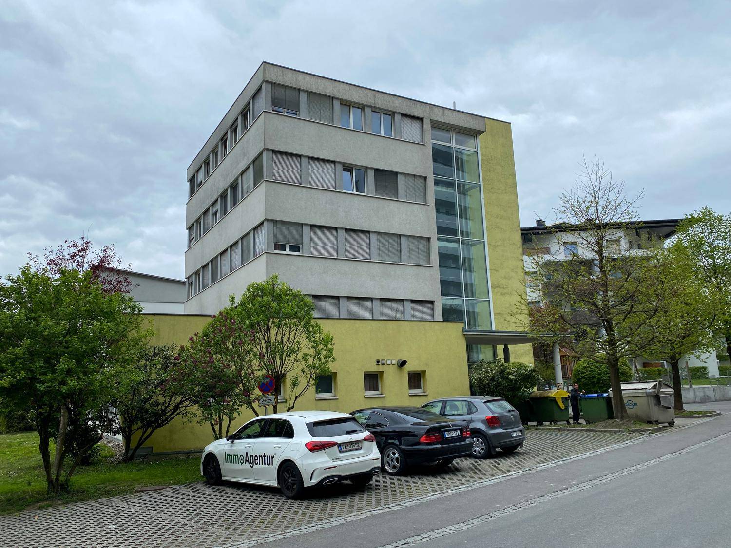 Ansicht des modernen Wohngebäudes von der Seite mit Parkplätzen und einem gläsernen Aufzugsschacht.