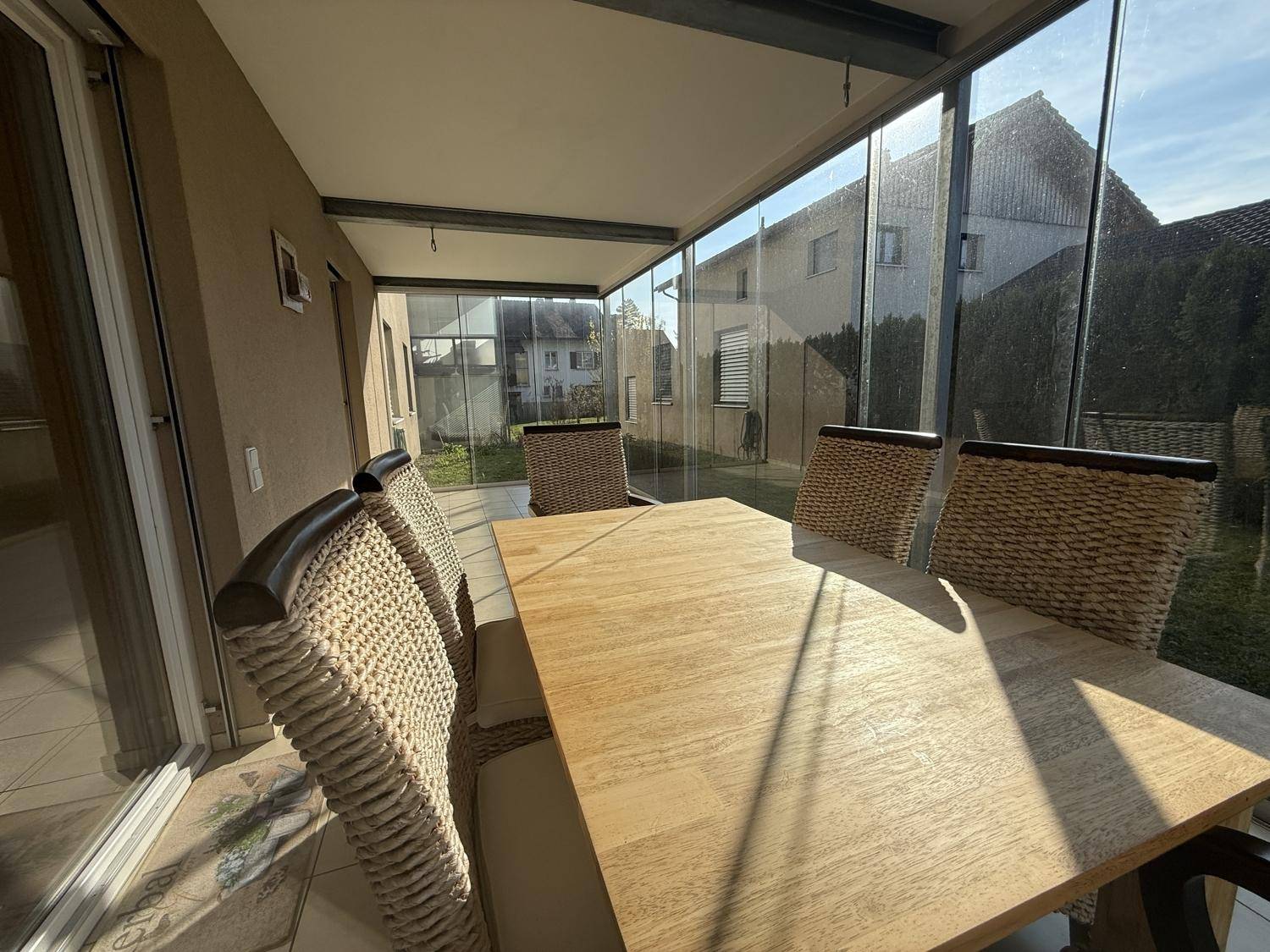 Essbereich auf der verglasten Terrasse mit Rattan-Stühlen und Holztisch, umgeben von Glaswänden.