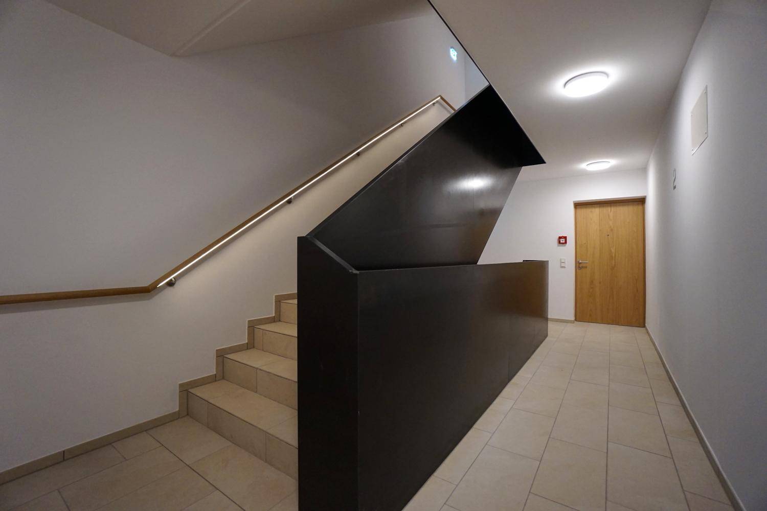 Modernes Treppenhaus mit elegantem Geländer und Fliesenboden, führt zu den Wohnungen.