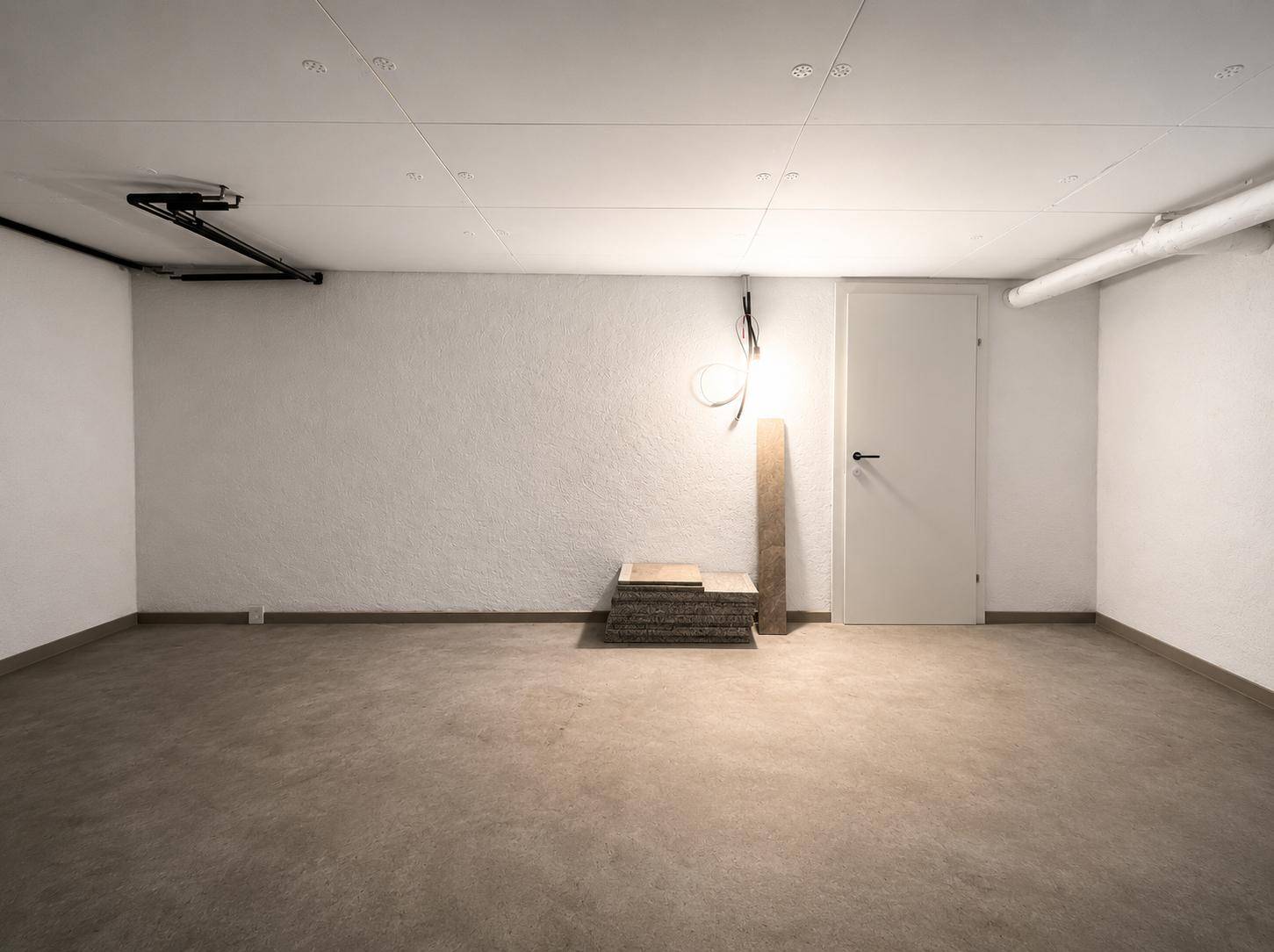 Leerer Raum mit weißen Wänden, Betonboden und sichtbaren Installationen, ideal als Keller oder Abstellraum.