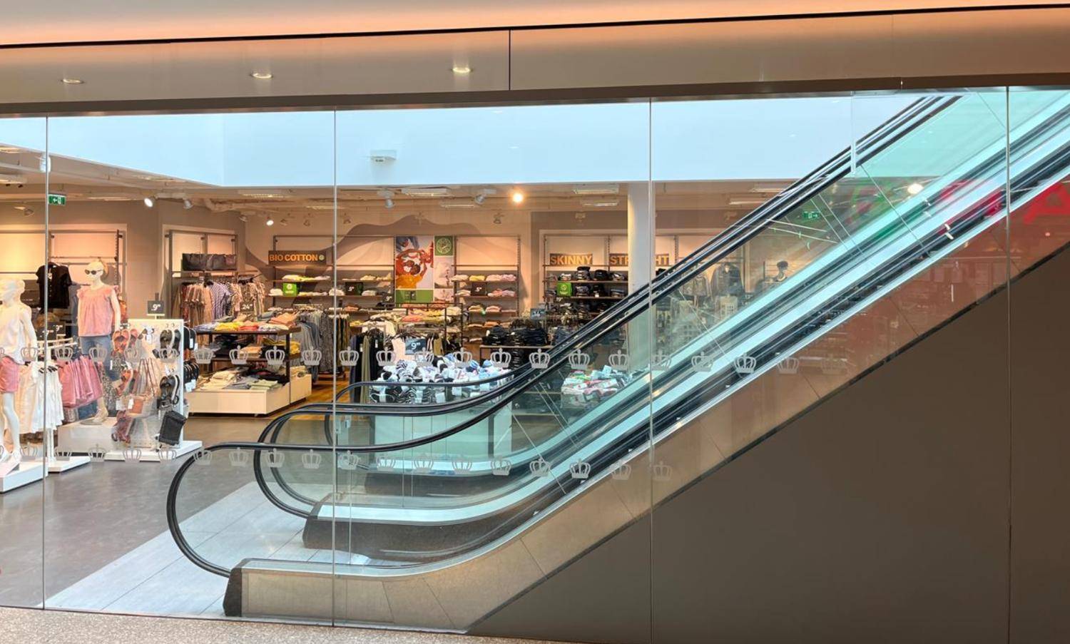 Innenansicht eines modernen Geschäfts mit Rolltreppe und Kleiderauslagen, hell beleuchtet.