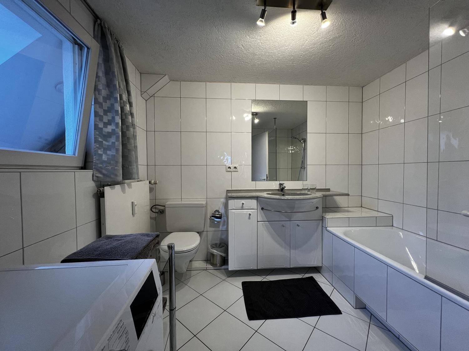 Gefliestes Badezimmer mit Badewanne, Waschbecken, WC und Waschmaschine, inklusive Fenster.